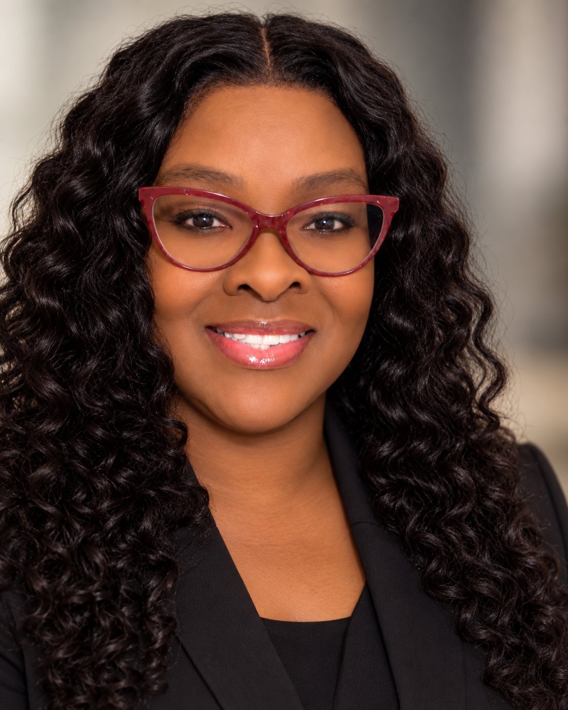 Close-up portrait of a woman with long, curly black hair, wearing red glasses, and a black blazer, smiling.