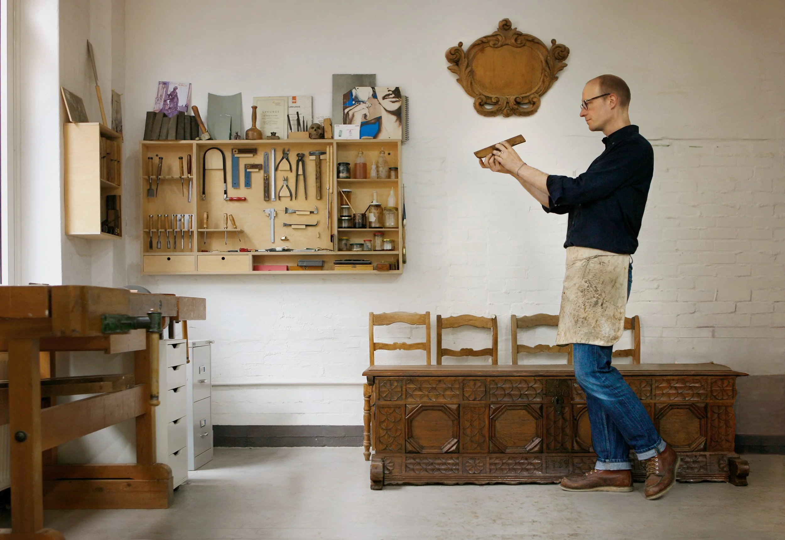 Ein Mann steht in einer Werkstatt, trägt eine Lederhose und hält ein Werkzeug in der Hand. Im Hintergrund sind Werkzeuge an einer Wand befestigt, sowie Bücher, Skulpturen und andere Gegenstände auf einem Regal. Der Raum hat weiße Wände und eine Holzbank.