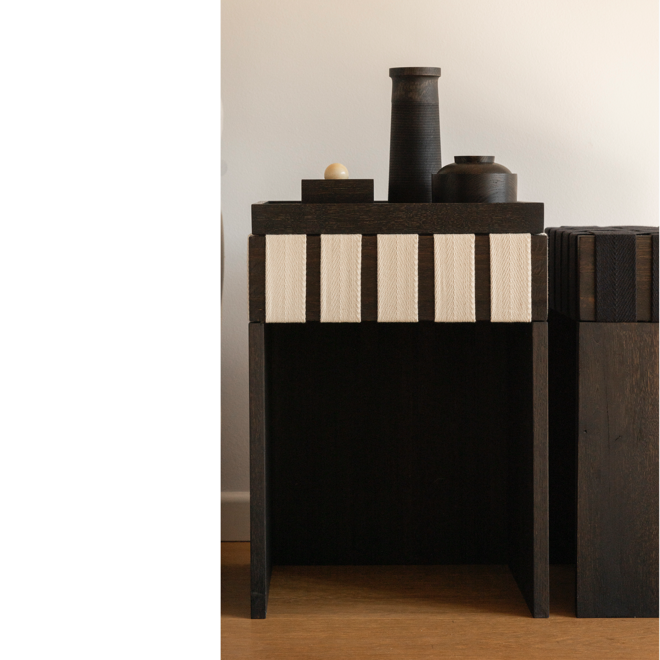 Dunkle Holz-Vase, eine kleine helle Kugel auf einem Ständer, dunkler Deckel, alles auf einem dunklen Holztisch gegen weiße Wand.