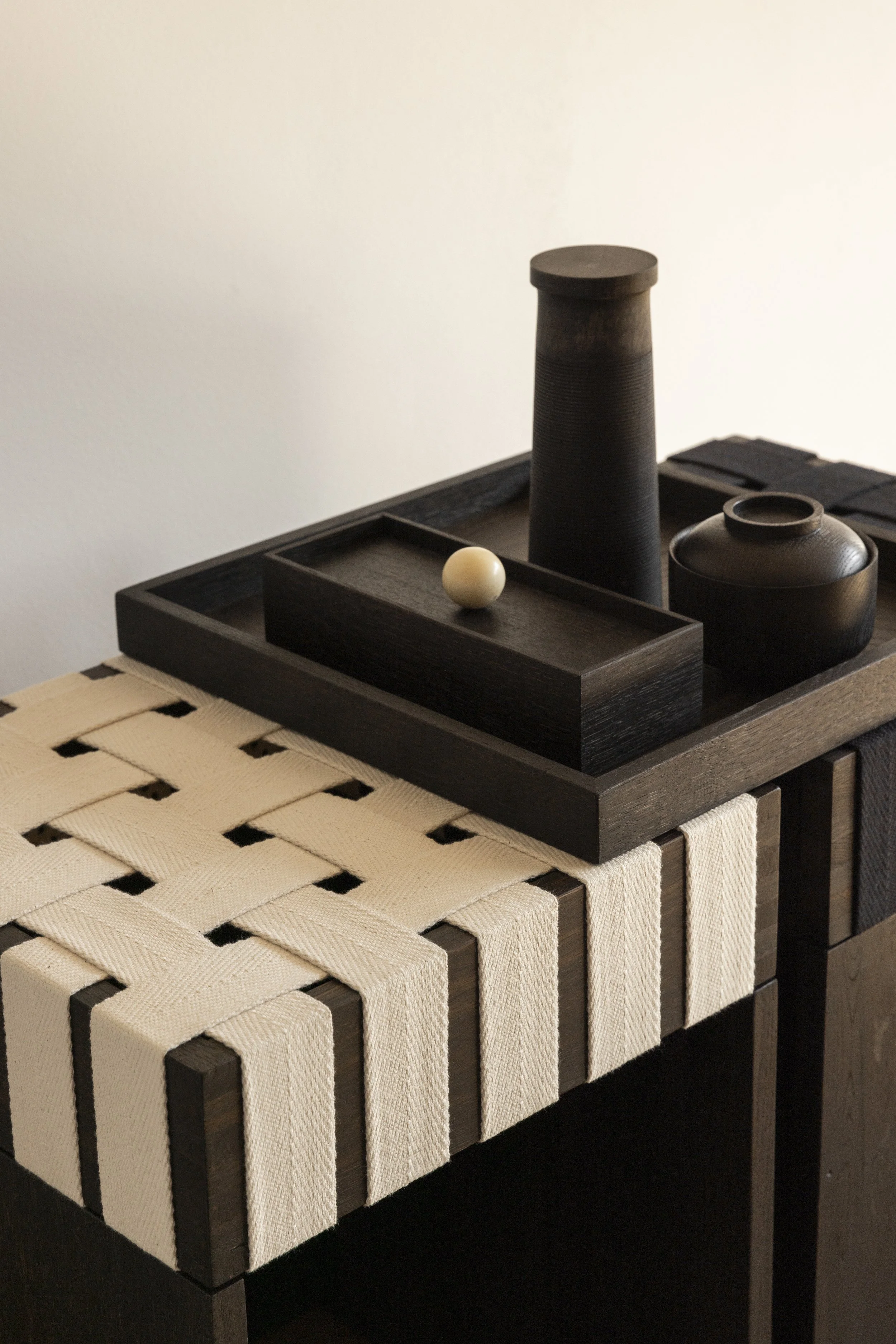 Dunkle Holztabelle mit schwarzer Ablagefläche, auf der sich eine dunkle Vase, eine schwarze Schachtel und eine kleine weiße Kugel befinden. Die Tabelle ist auf einem beige geflochtenen Untersetzer an einer weißen Wand.