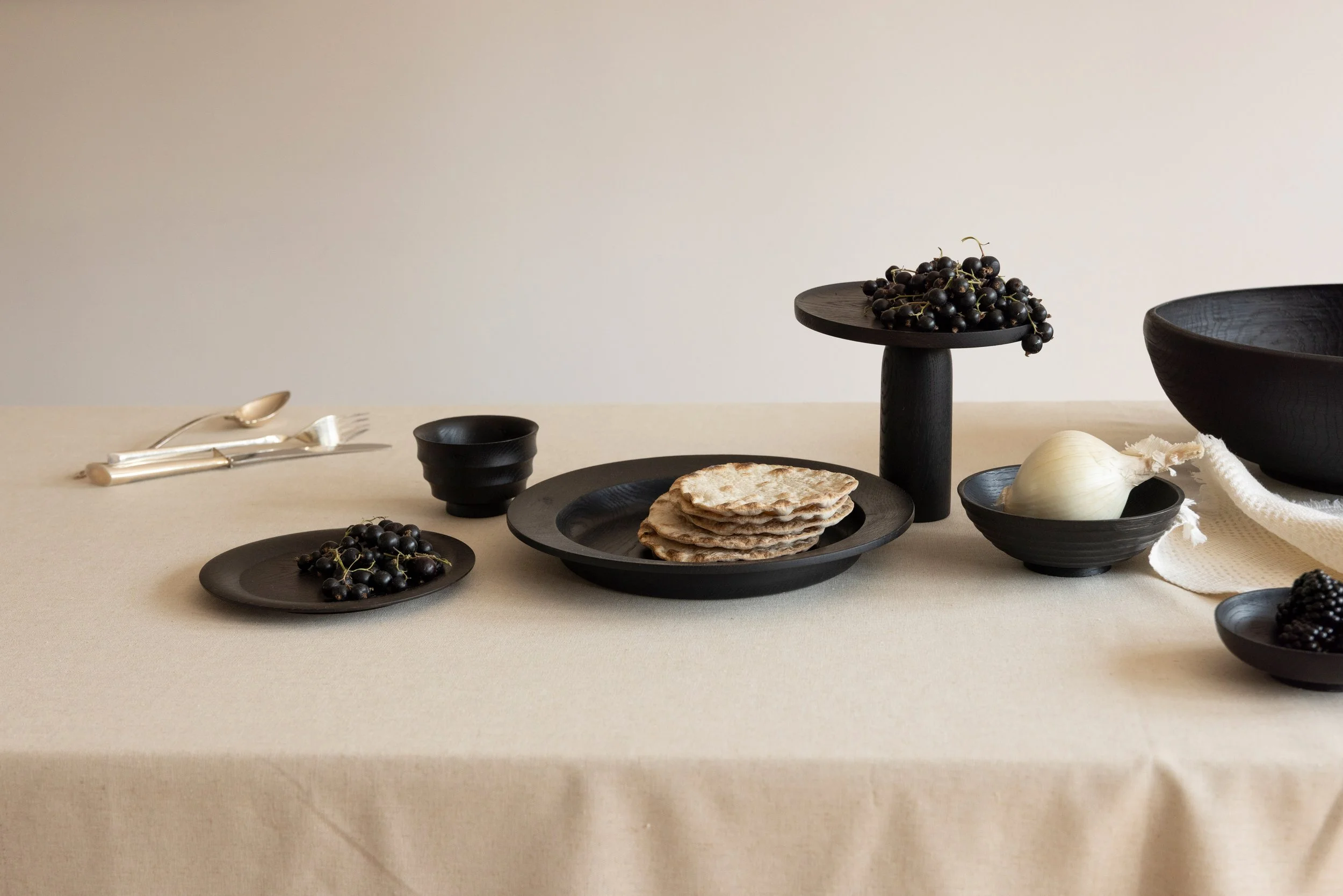 Tisch mit schwarzen Tellern, einem schwarzen Schüssel, einer schwarzen Kerze auf einem schwarzen Ständer, frischen Trauben, Fladenbrot und einer Zwiebel auf einer weißen Stoffserviette, mit silbernem Besteck auf der linken Seite.
