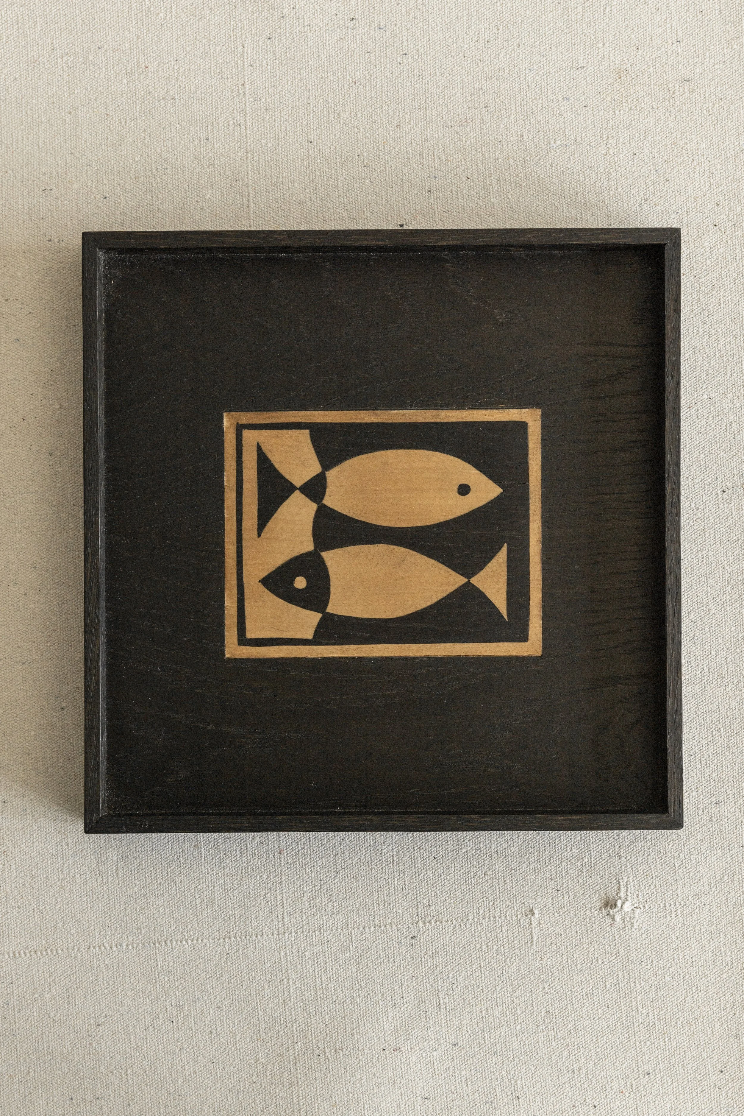 Ein schwarzer Bilderrahmen mit einer Illustration von drei goldfarbenen Fischen auf hellbraunem Hintergrund, aufgehängt an einer Wand.