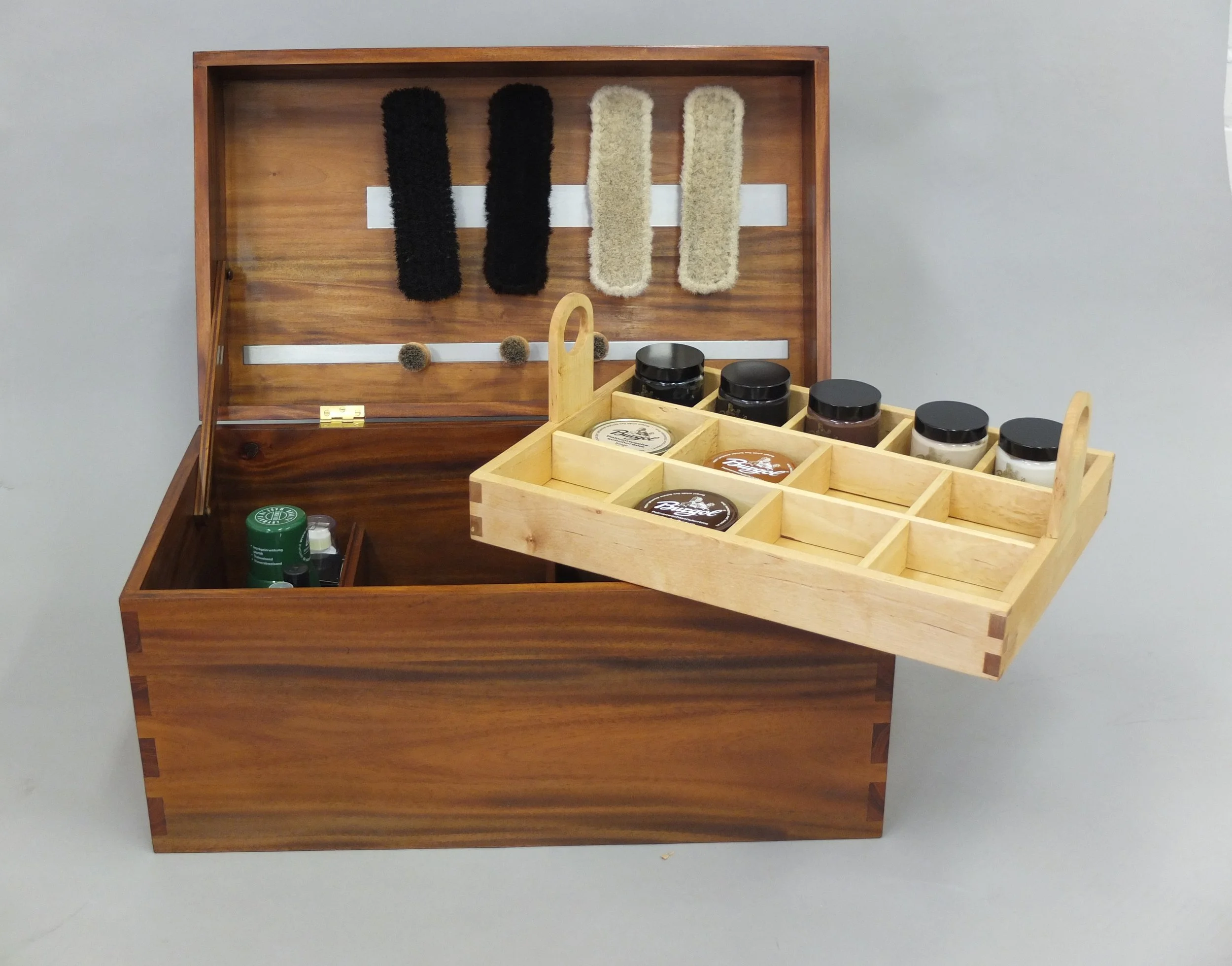 Ein Holzkästchen mit verschiedenen kleinen Gegenständen, darunter ein grüner Behälter, einige Gläser mit schwarzen Deckeln und eine Aufbewahrungsbox mit mehreren Fächern, die Cremes und andere Produkte enthalten. Im Hintergrund eine Wand mit Haken, a