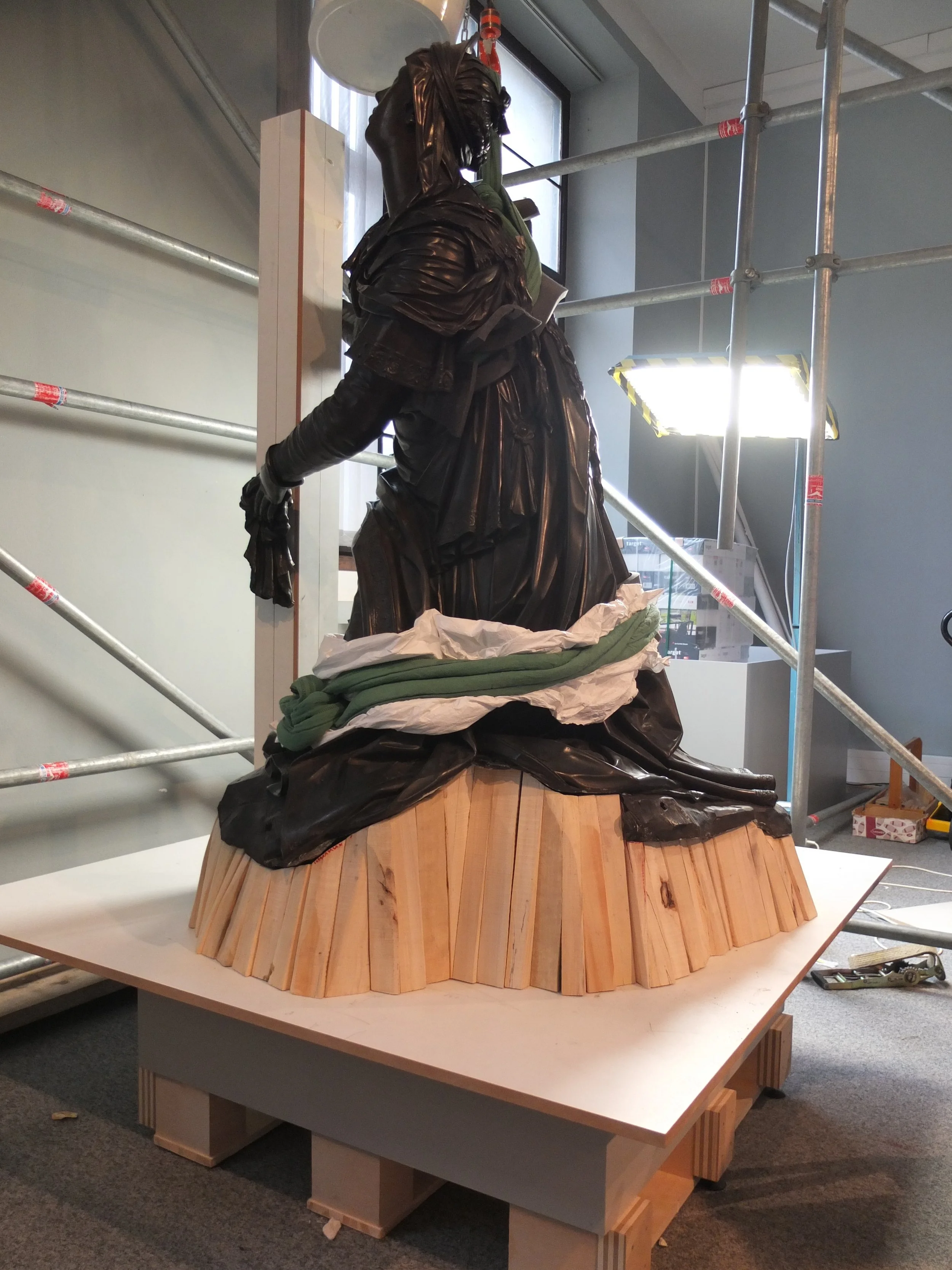 Eine Statue eines Menschen in dunklen Kleidern auf einem Holzpodest, umgeben von Gerüsten und Baumaterialien in einem Raum mit großen Fenstern.