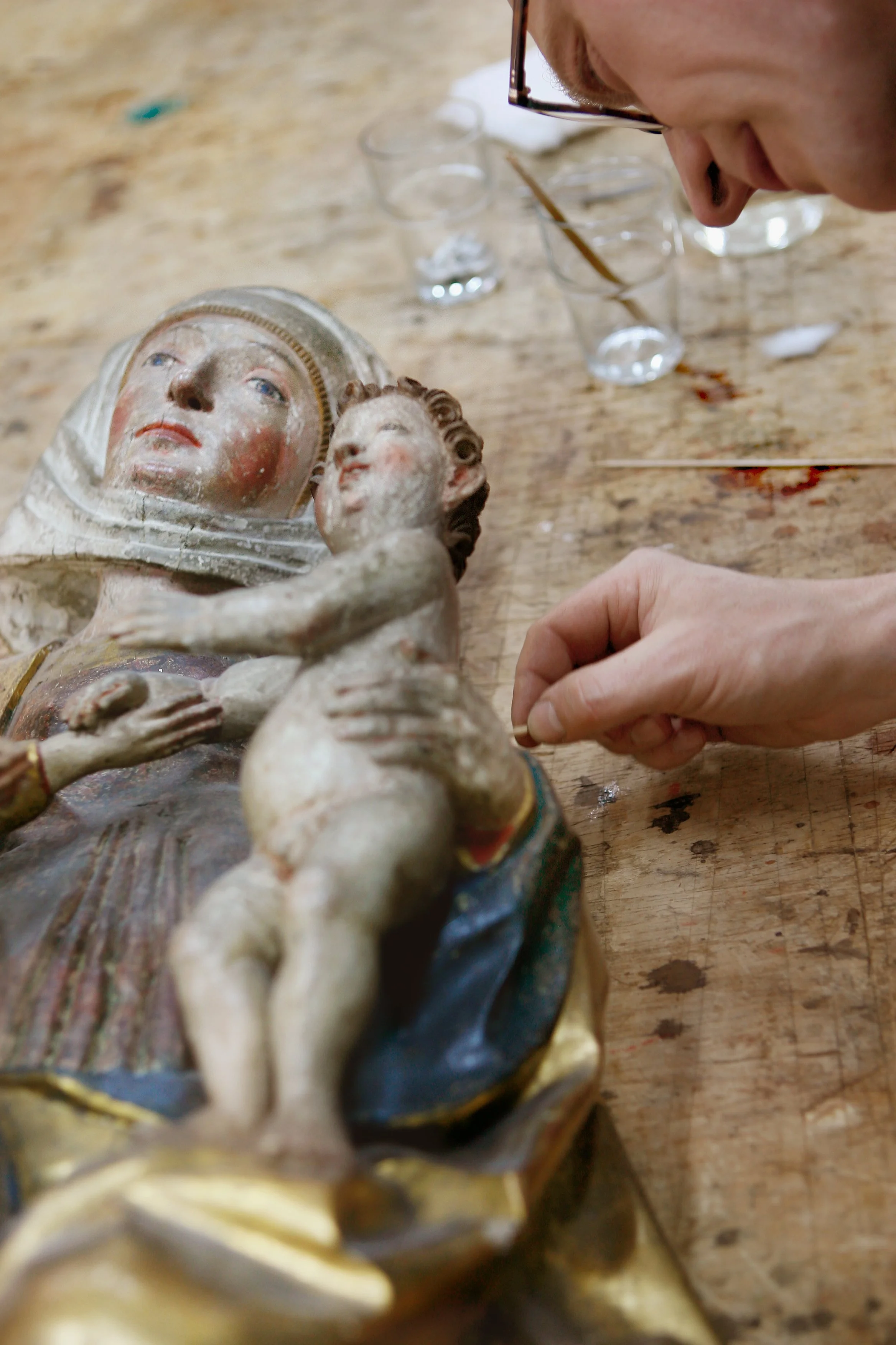Ein Mensch schaut auf eine antike, beschädigte Statue, auf einem Holztisch mit mehreren Gläsern im Hintergrund.
