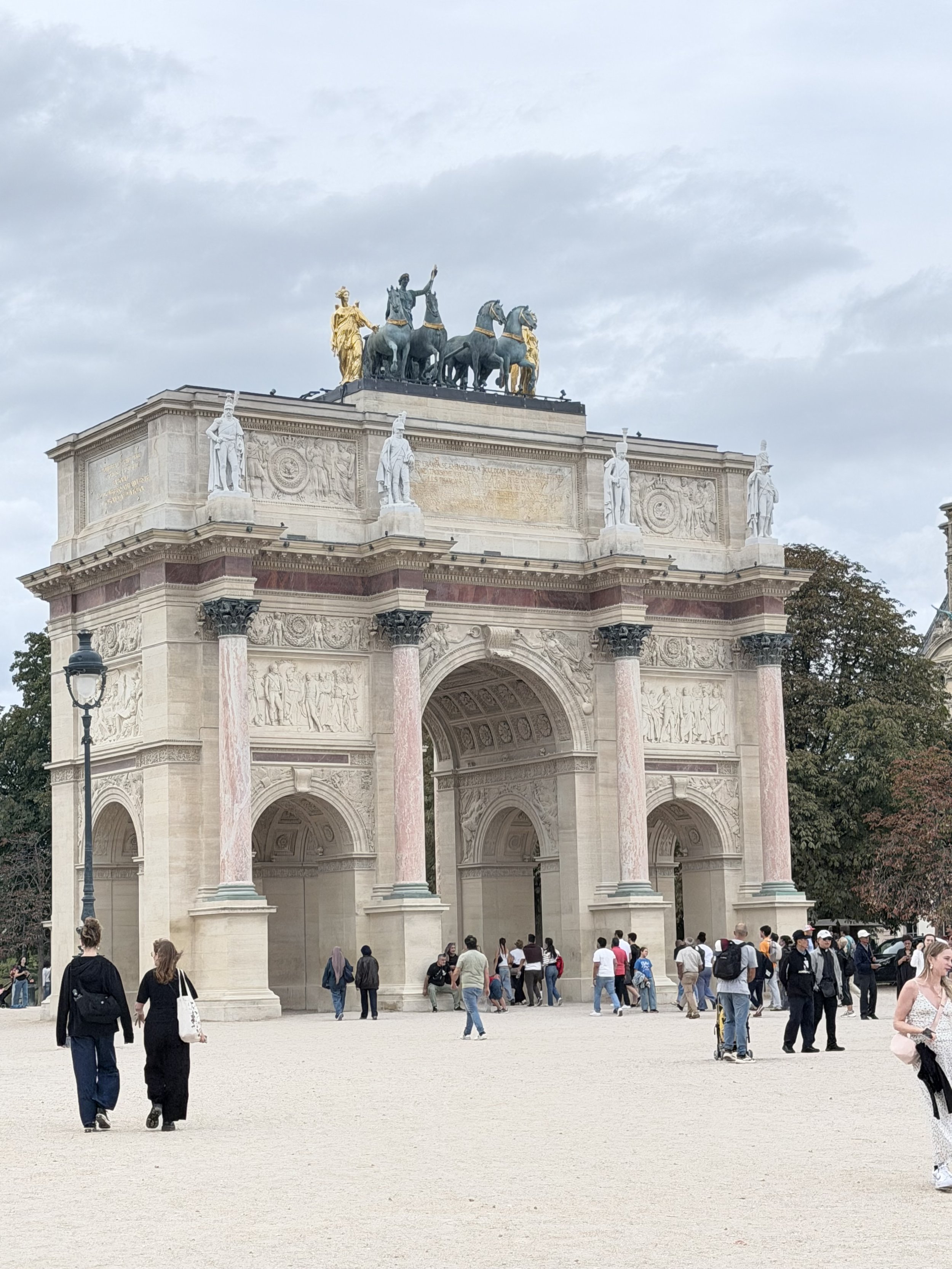 People walking and gathering around a large, ornate triumphal arch with sculptures and a golden chariot on top, under a cloudy sky.