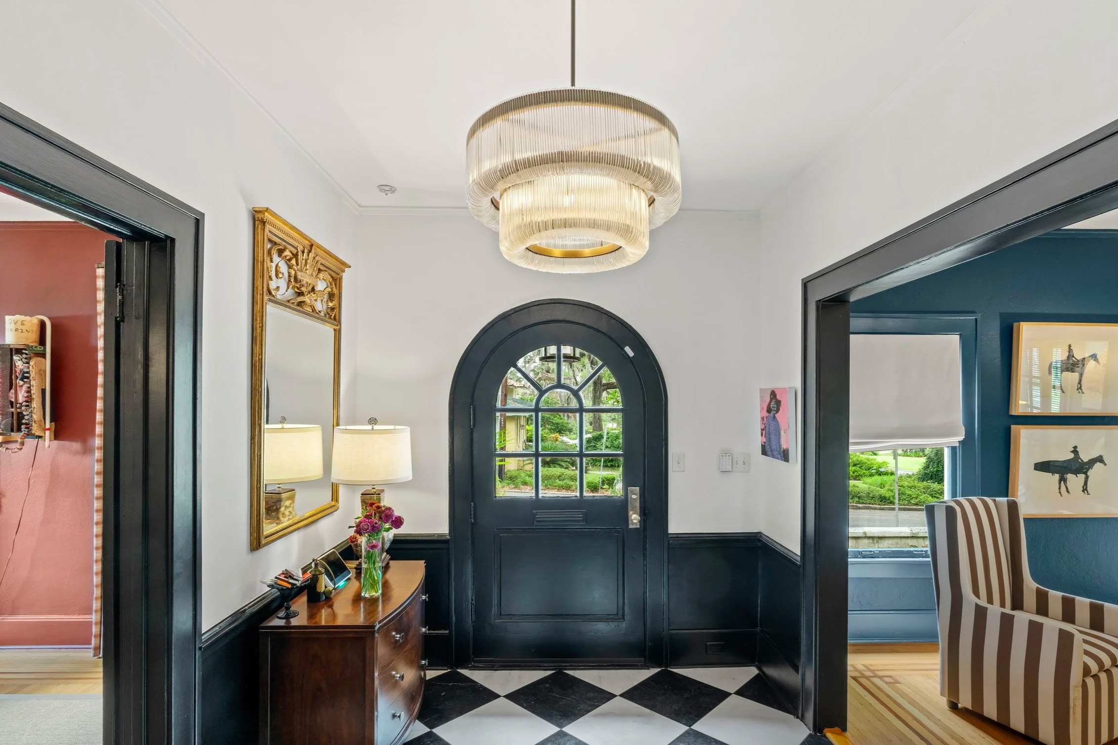 An entryway with a dark door and a large window. There is a chandelier hanging from the ceiling, and a mirror with a gold ornate frame on the wall. A small table with flowers and books is below the mirror, and an armchair is visible to the right.