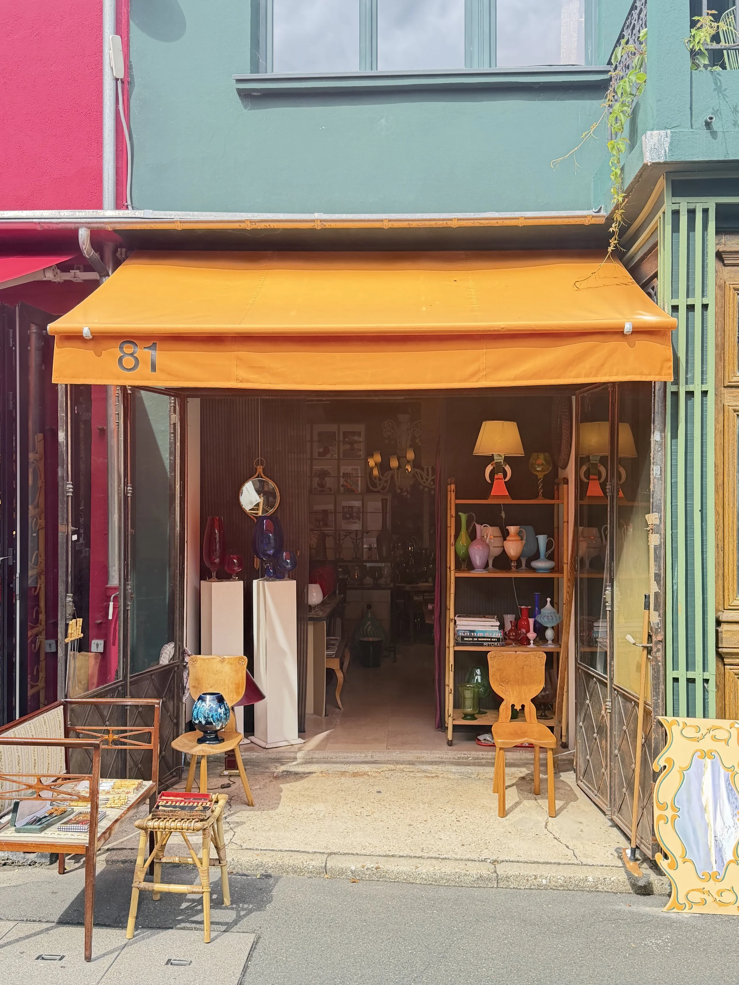 View of a storefront with a yellow awning displaying the number 81. The store is open, showing shelves with colorful vases and decorative items inside. Outside, there are small tables and chairs, and the street pavement with a sidewalk is visible.
