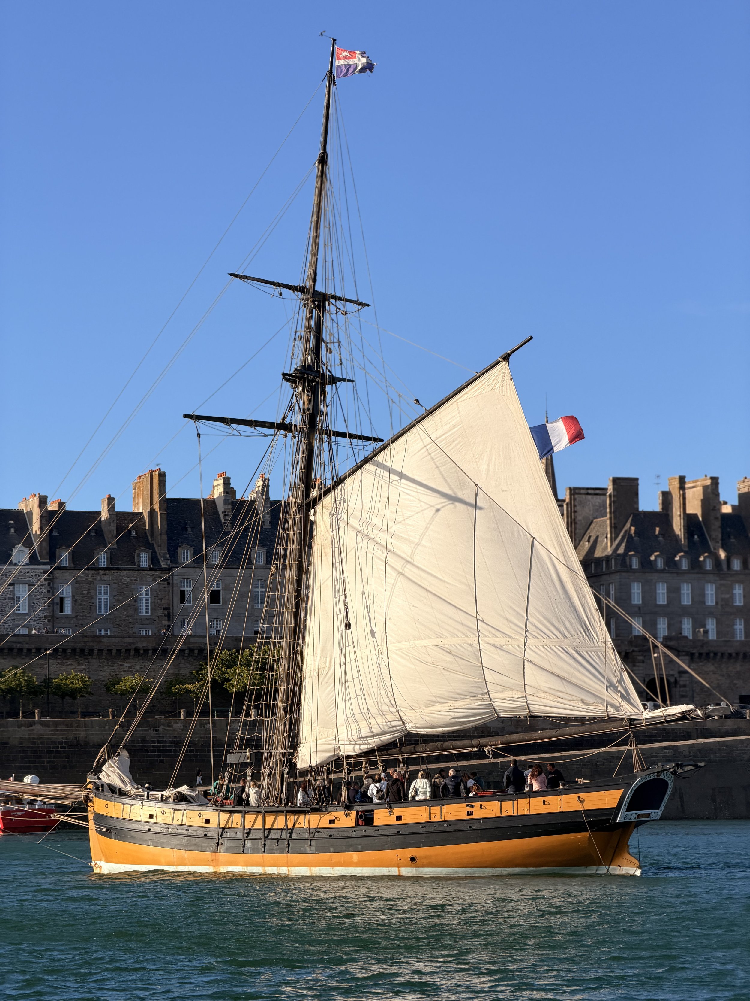 A large historic sailing ship with beige sails, a yellow and black hull, and the French flag, docked along a city harbor with buildings and people on the waterfront.