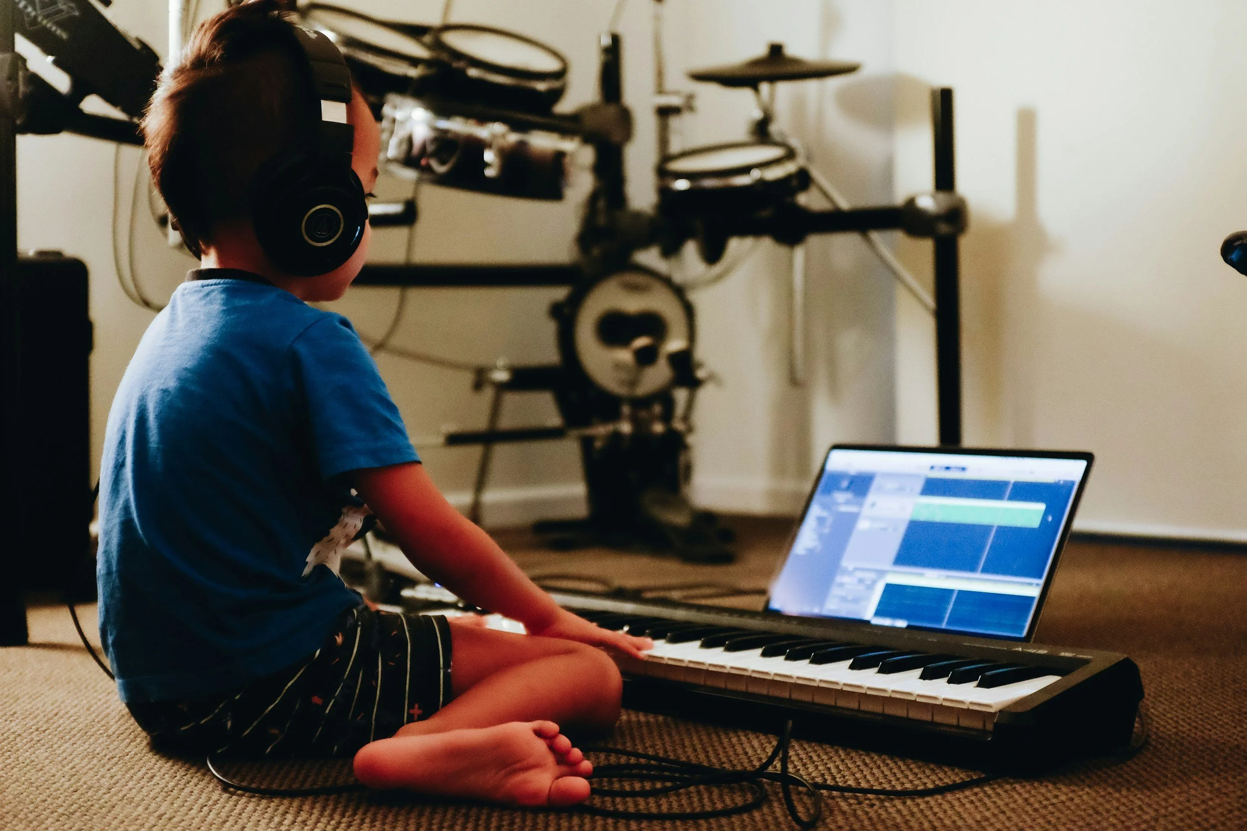 Niño sentado en el suelo tocando un piano digital con auriculares, frente a una computadora portátil con programa de edición musical, en una habitación con equipo de instrumentos musicales en el fondo.
