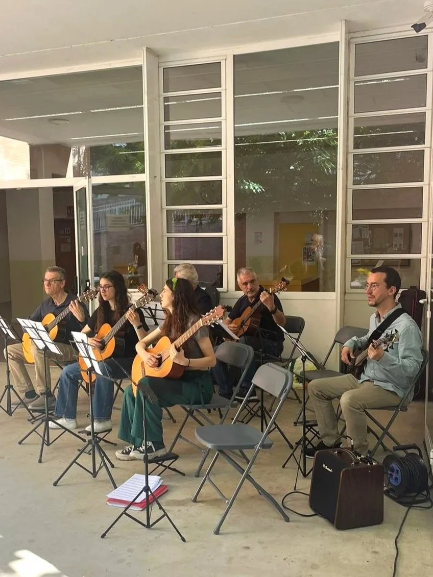 Grupo de seis personas tocando guitarras en un espacio interior con ventanas grandes y notas musicales en partituras en los soportes.