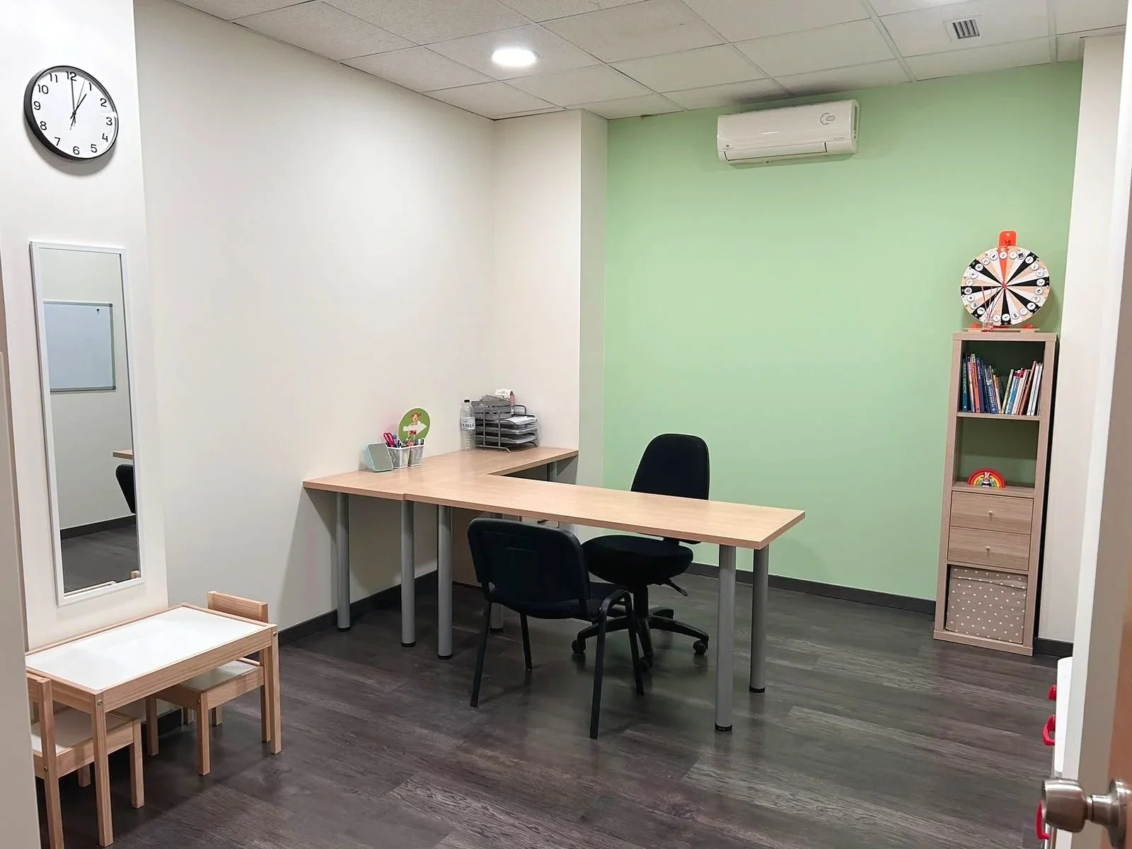 Oficina con muro verde y blanca, mesa en forma de L, dos sillas negras, estantería con libros, en la pared reloj y aire acondicionado, en el piso madera oscura.
