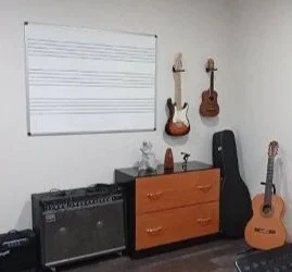 Sala de música amb 3 guitarres, un amplificador, un moble amb tres calaixs, i una pissarra amb pentagrames sobre una paret blanca.