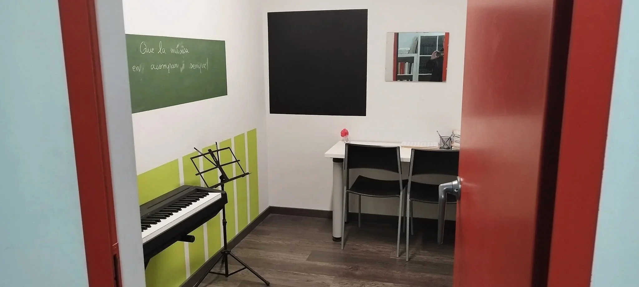 Sala de clases pequeña con un teclado eléctrico, una mesa con dos sillas, un espejo y un pizarrón verde con escritura en español.