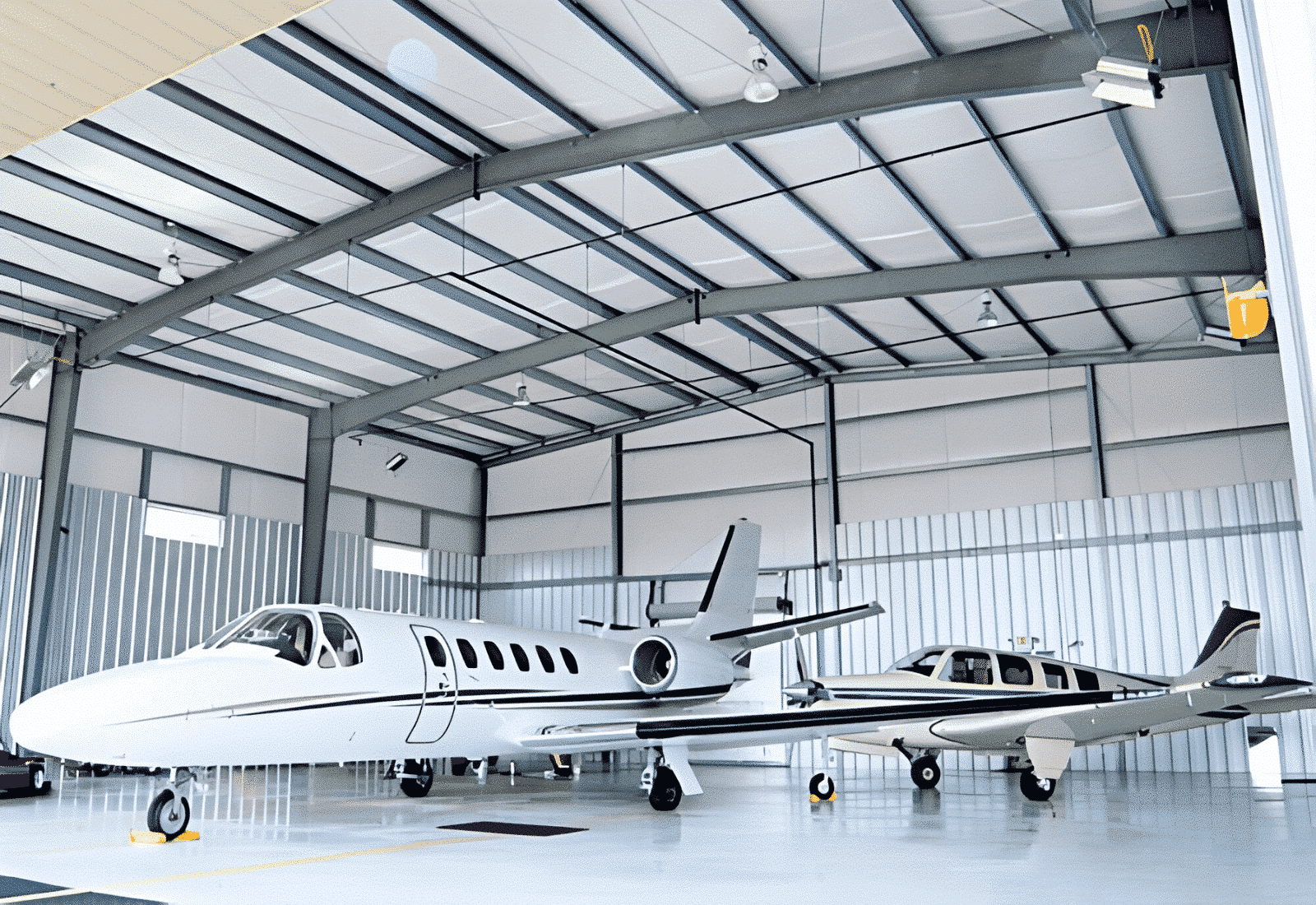 Inside a spacious aircraft hangar with a high ceiling and metal walls, featuring two private jets parked side by side on a smooth concrete floor.