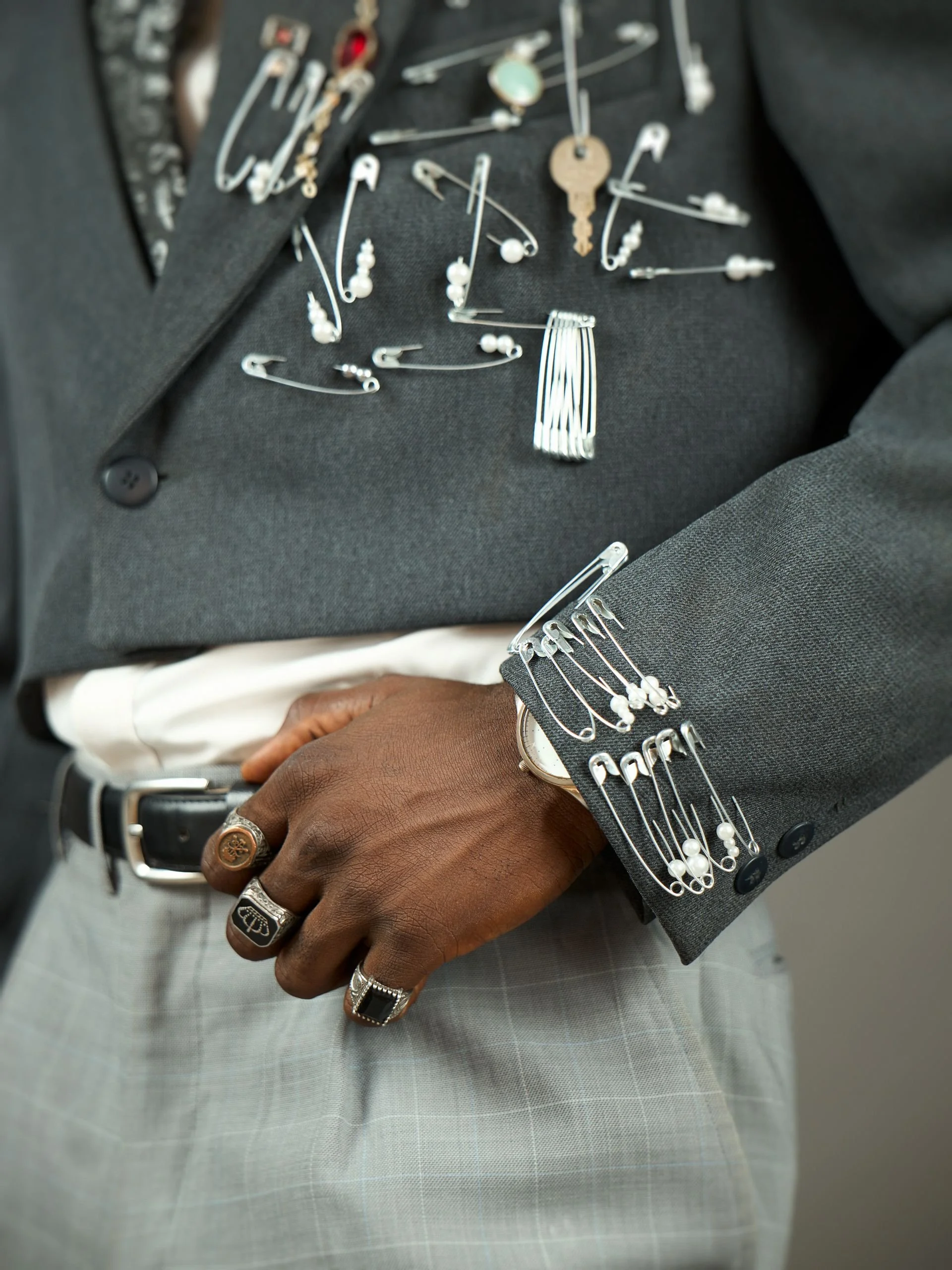 Close-up of a person's right hand showing several signature rings, a large ring, and a bracelet. The person is wearing a gray checked suit with cufflinks. On their wrist is a watch and a bracelet made of safety pins. The suit sleeve has safety pins with pearls attached, and the background shows a collection of safety pins, sewing pins, and jewelry components on a black fabric surface.