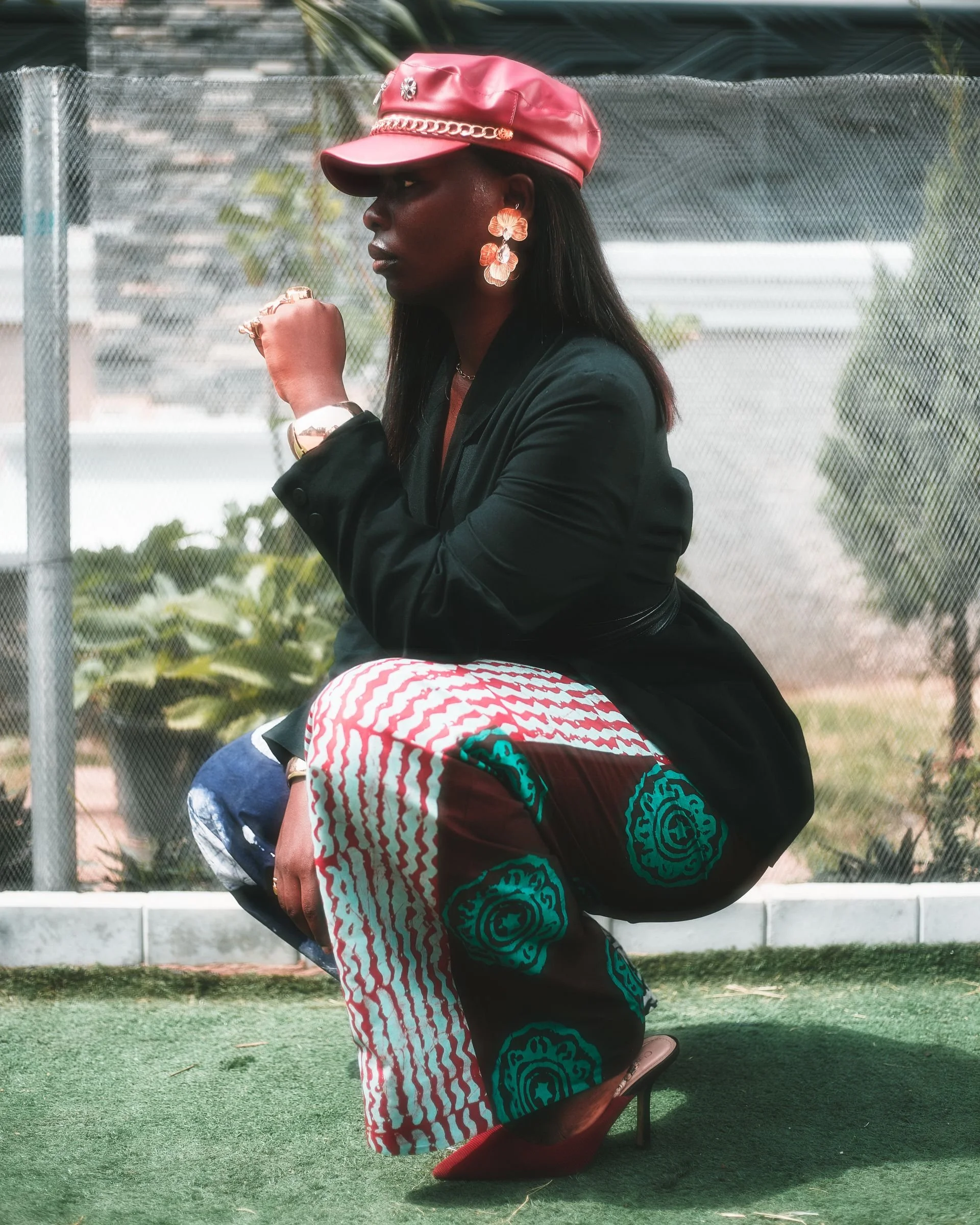 A woman with dark skin wearing a red leather cap with a chain accessory, large flower-shaped earrings, a black jacket, and patterned pants with red and white wavy lines and blue circular designs, crouches with her left fist near her face, outdoors in front of a chain-link fence with greenery behind.