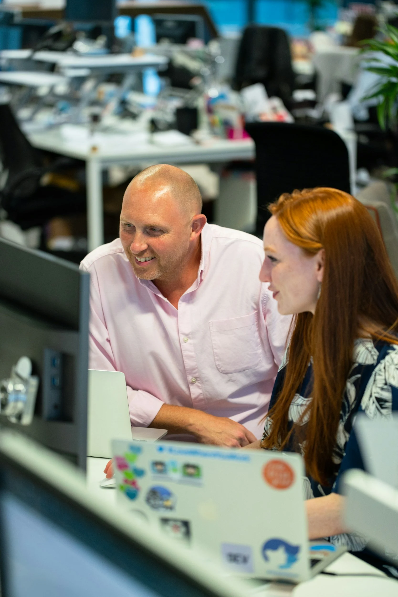 A man and a woman sitting at a desk, looking at a computer screen and smiling in an office environment.