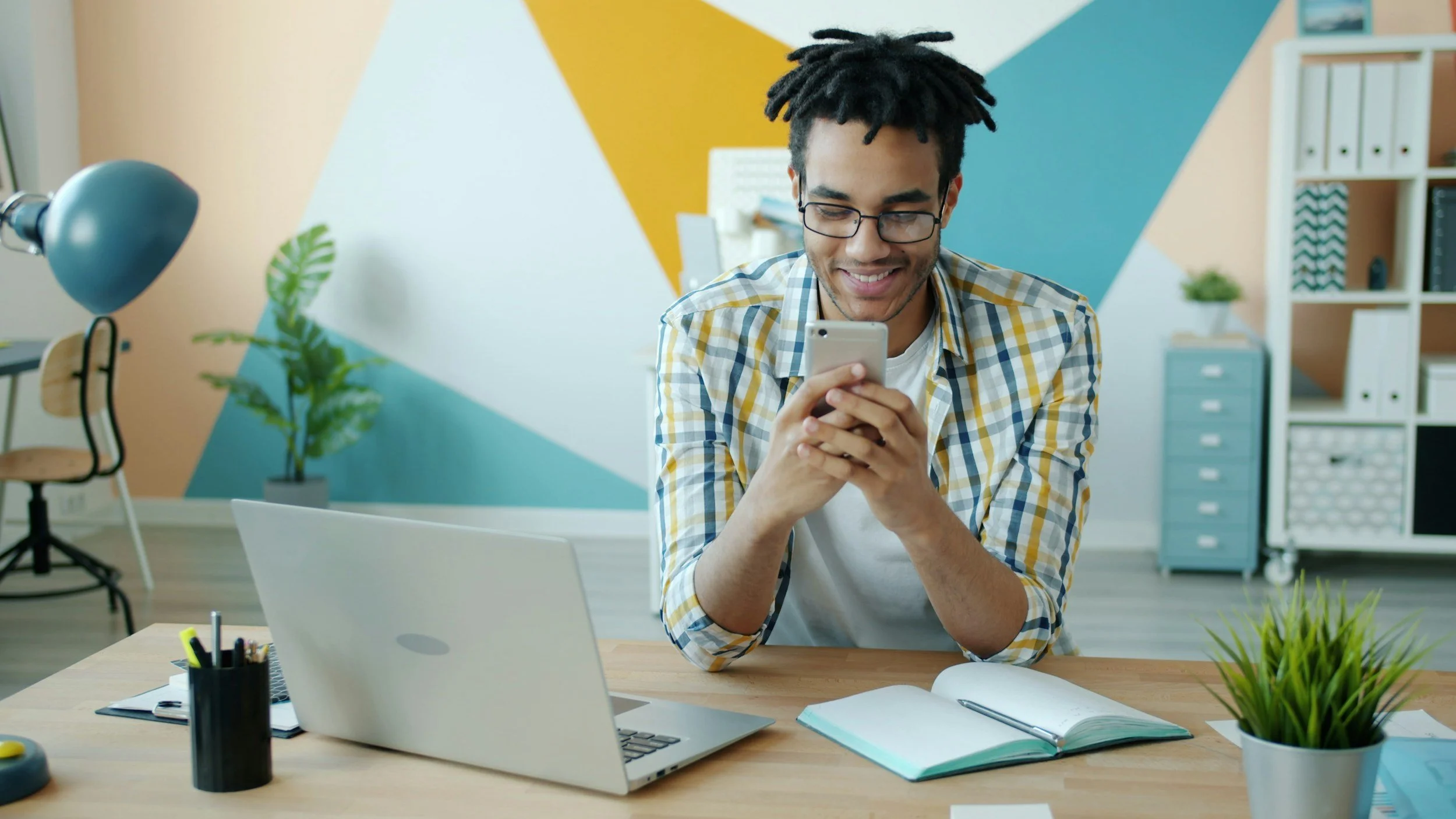 Young man with glasses smiling while looking at his smartphone in a modern office. Desk with open laptop, notebooks, and pens; abstract colorful wall, plants, and office furniture in the background.