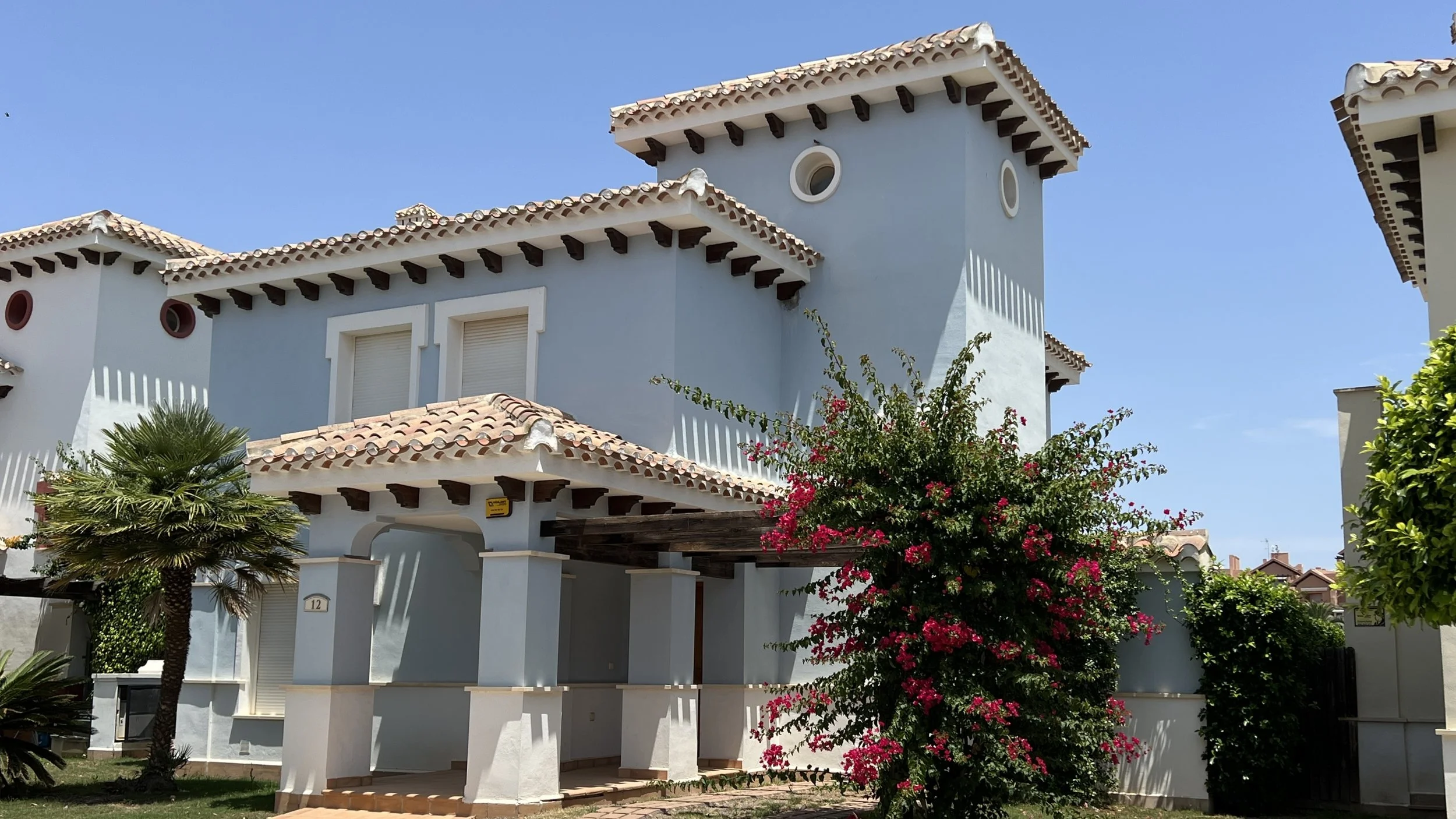 White Mediterranean-style house with red-tile roof, surrounded by greenery and flowering bushes, under a clear blue sky.