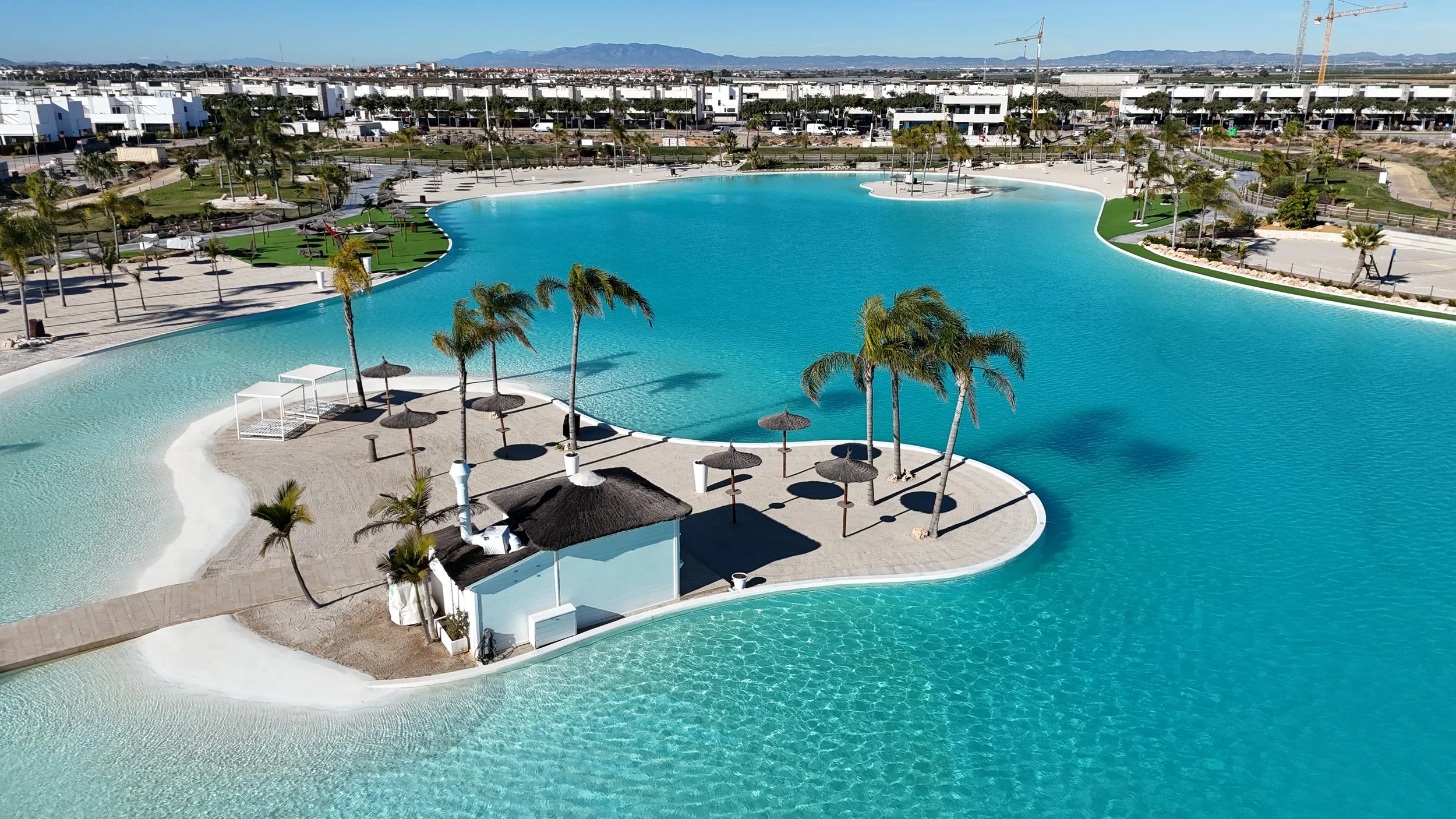 A large man-made lagoon with clear blue water, surrounded by palm trees, thatch umbrellas, and a sandy beach area. There are modern white buildings and construction cranes in the background, with mountains visible in the distance.