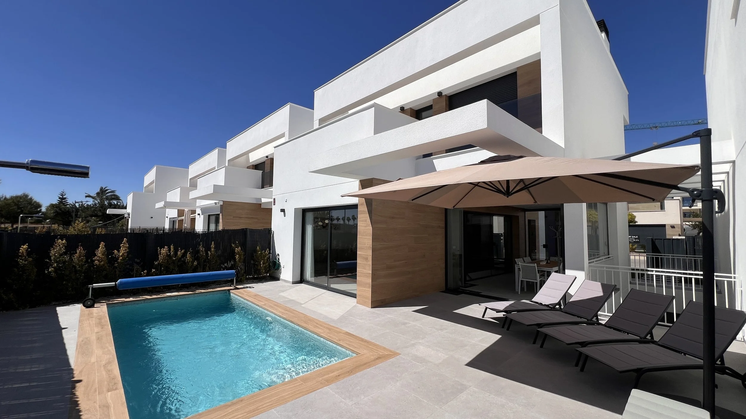 Modern white multi-story residential building with balconies, a backyard pool, outdoor lounge chairs, and umbrellas on a sunny day.