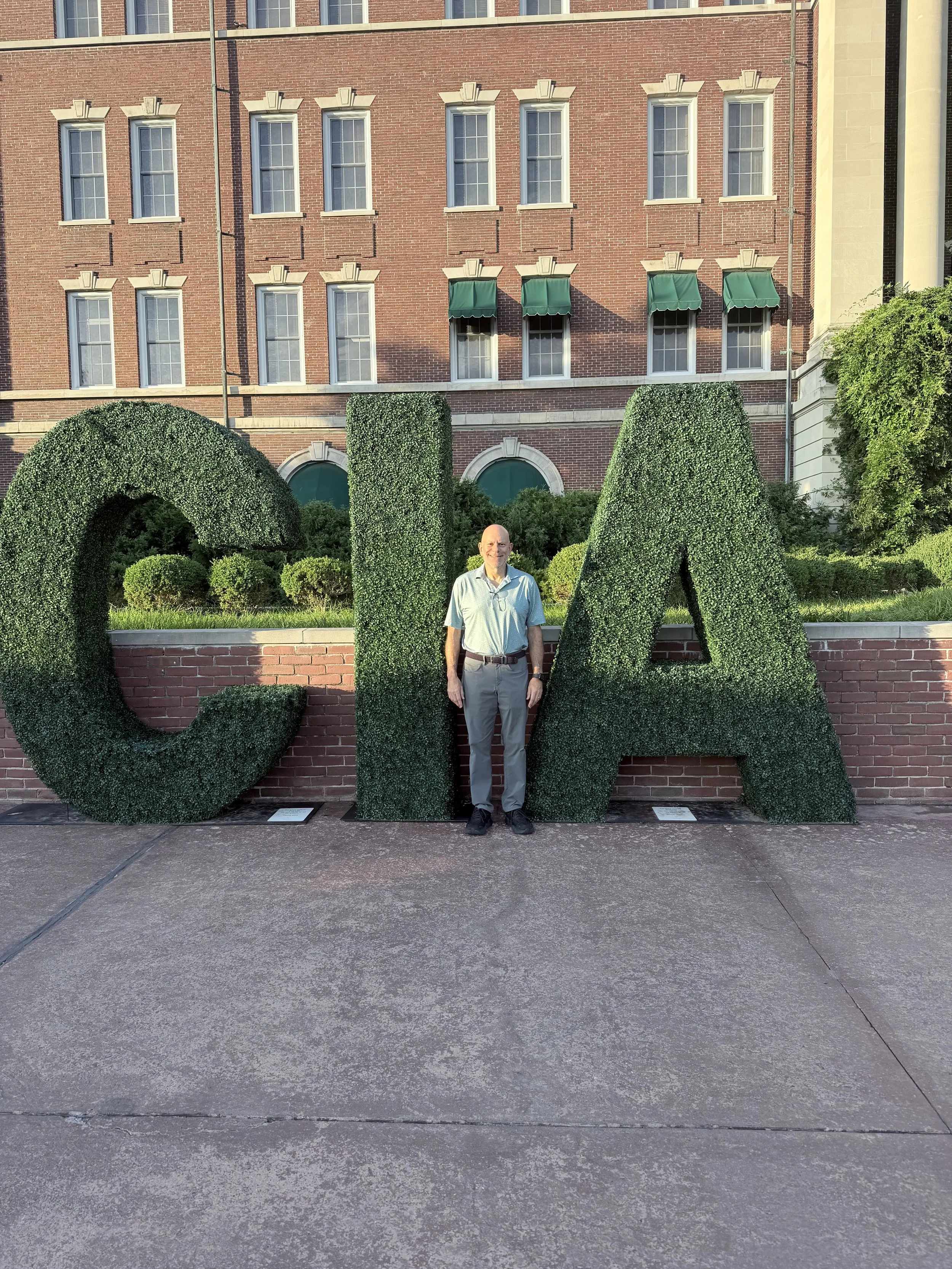 A man standing in front of large green topiary letters that spell out 'CIA' with a brick building in the background.