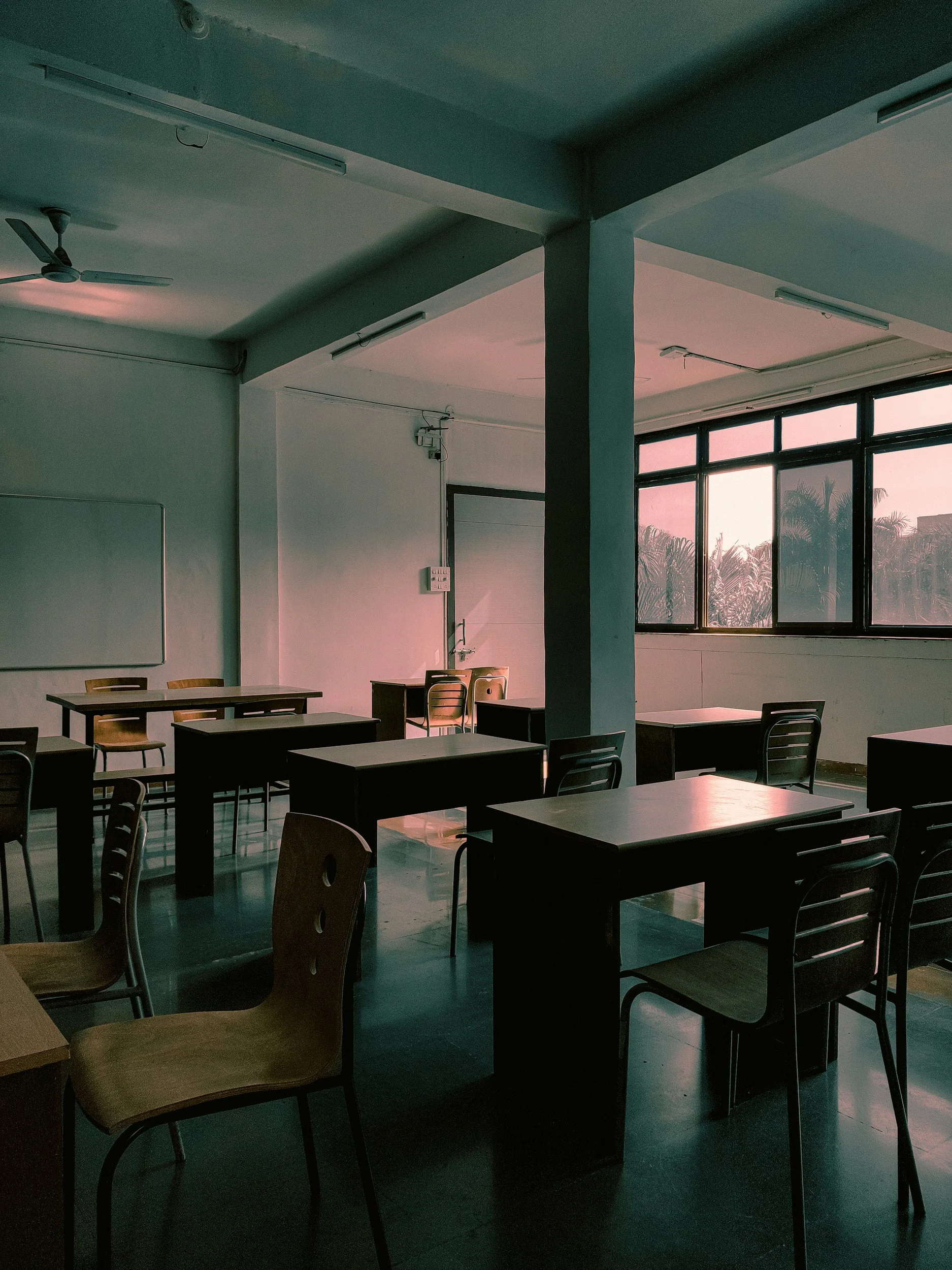 Sala de aula vazia com janelas grandes, várias mesas e cadeiras de madeira, parede branca, luminárias no teto e ventilador.