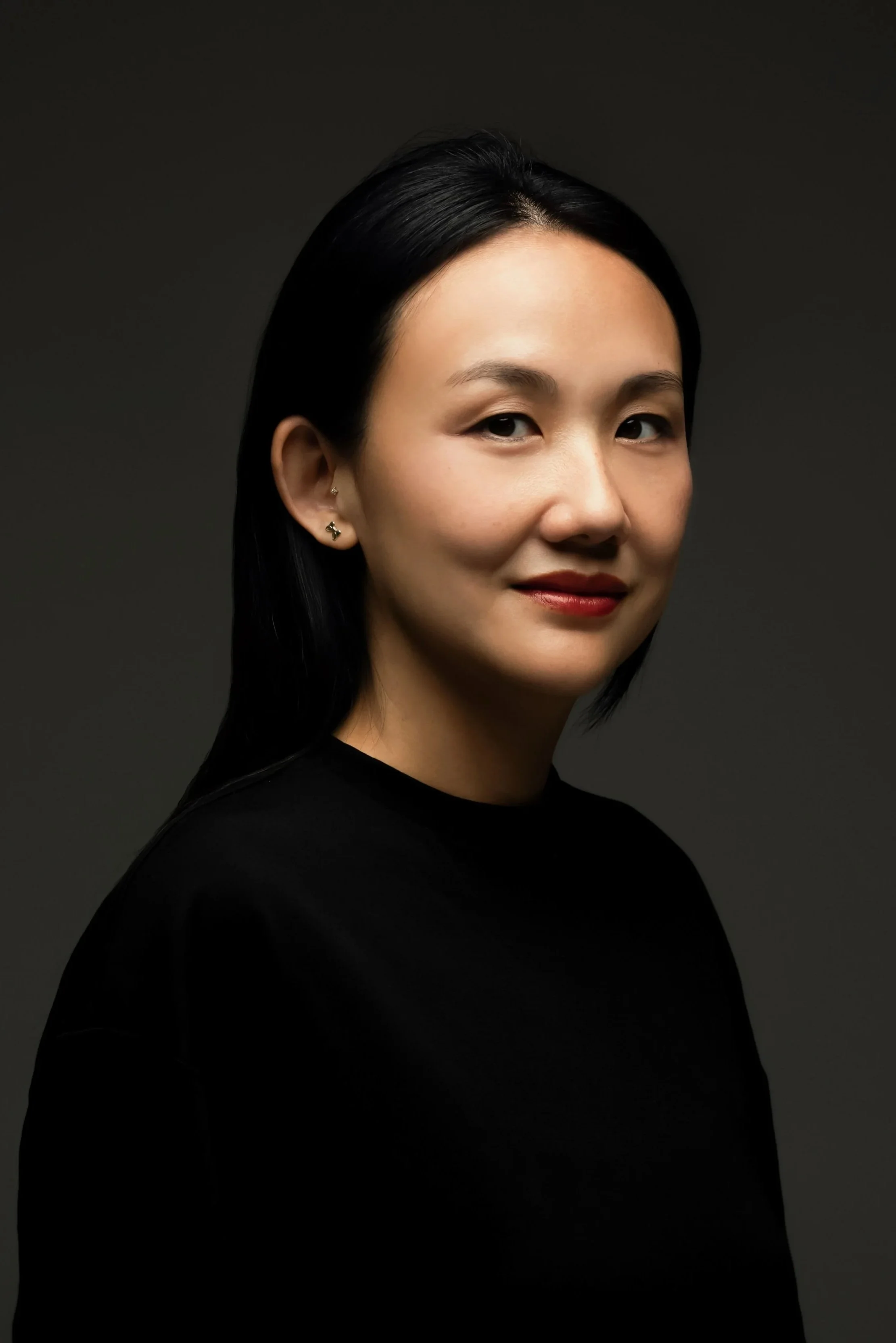 A woman with black hair, wearing a black top, is shown in a portrait against a dark background. She has small earrings and a subtle smile.