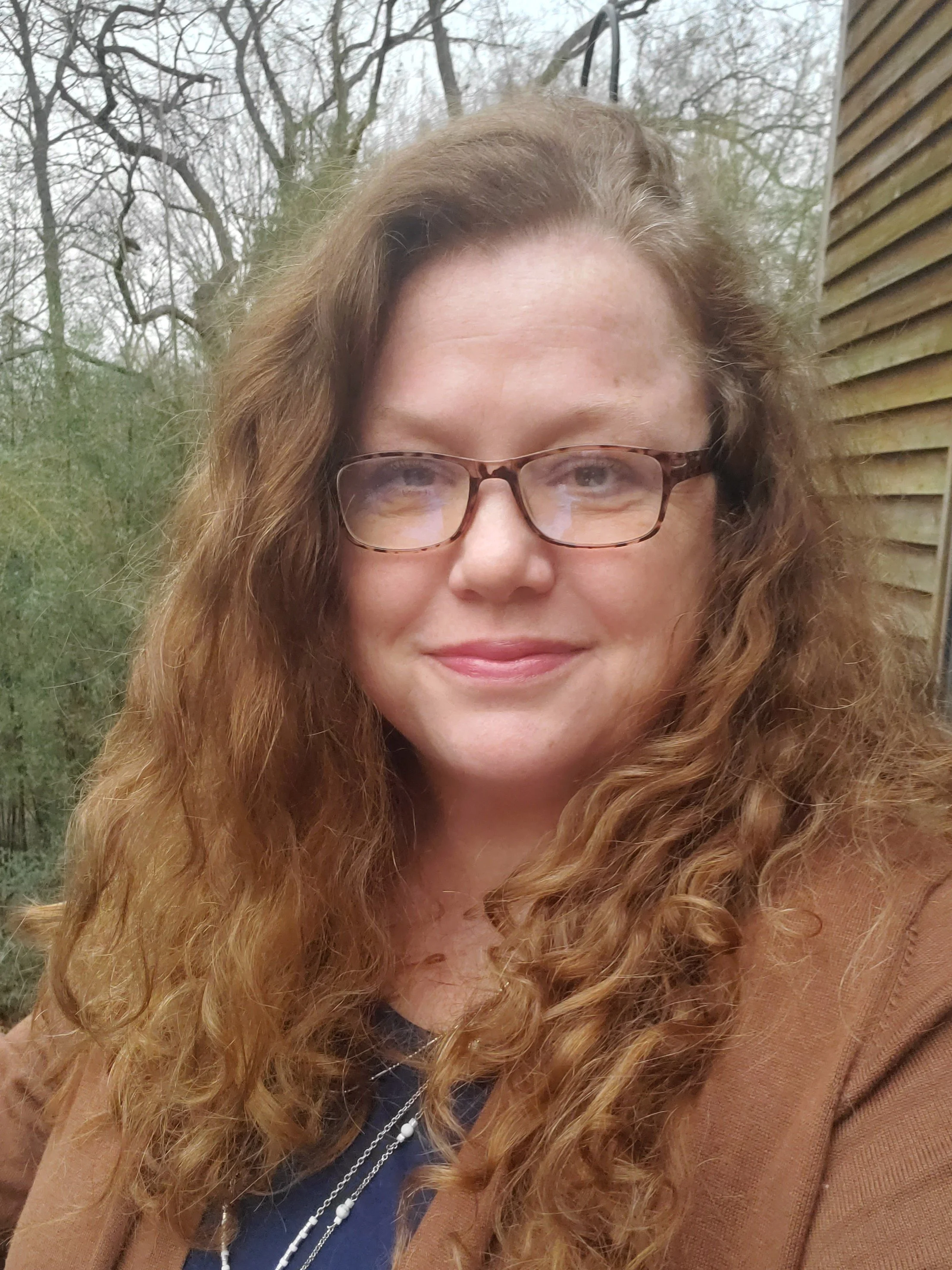 Close-up of a woman with long curly red hair and glasses, smiling outdoors with a wooden wall on the right and leafless trees in the background.