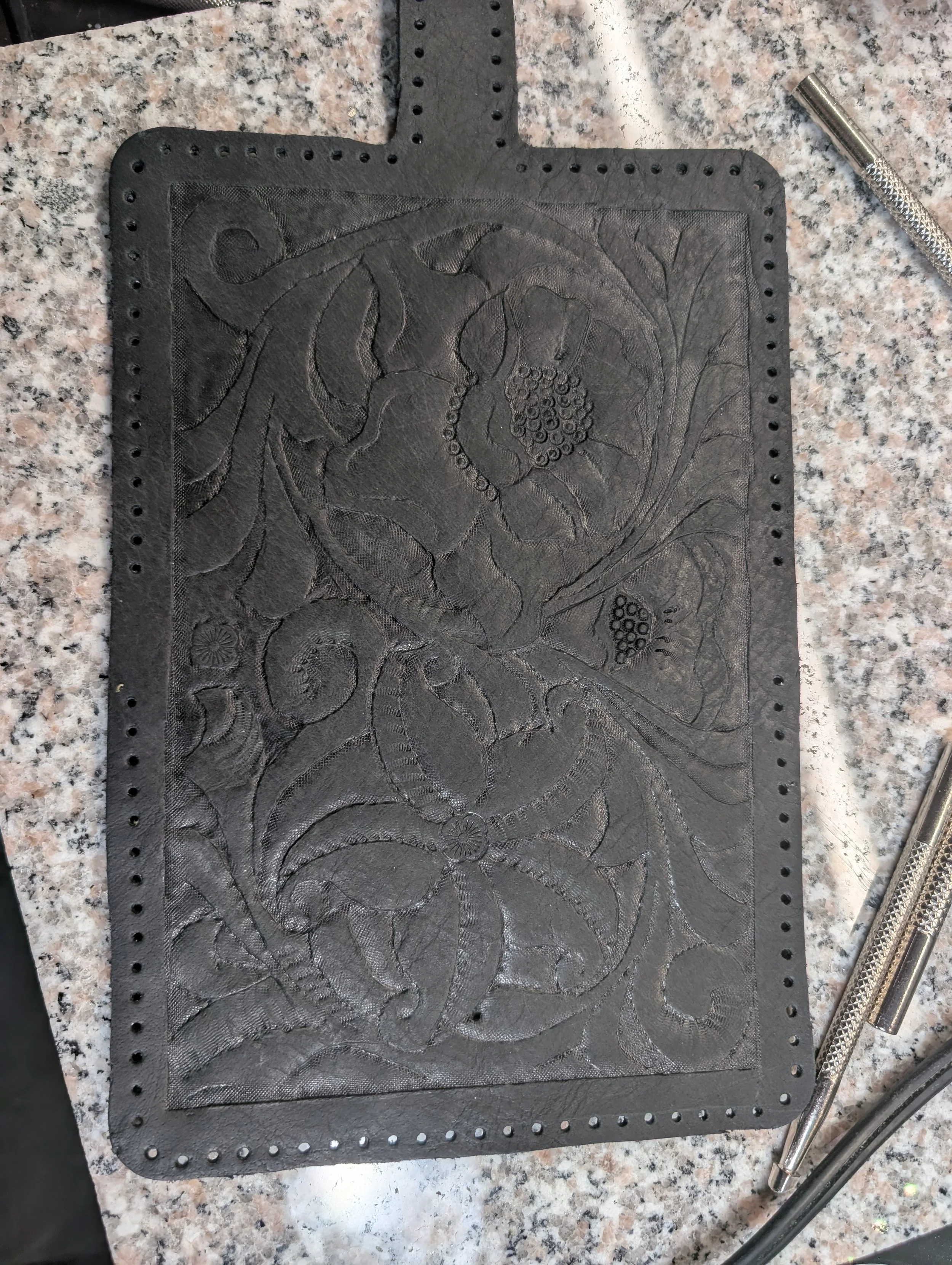 A black textured rubber mat with an engraved floral pattern, placed on a speckled granite countertop, with a silver metal tool on the right side.