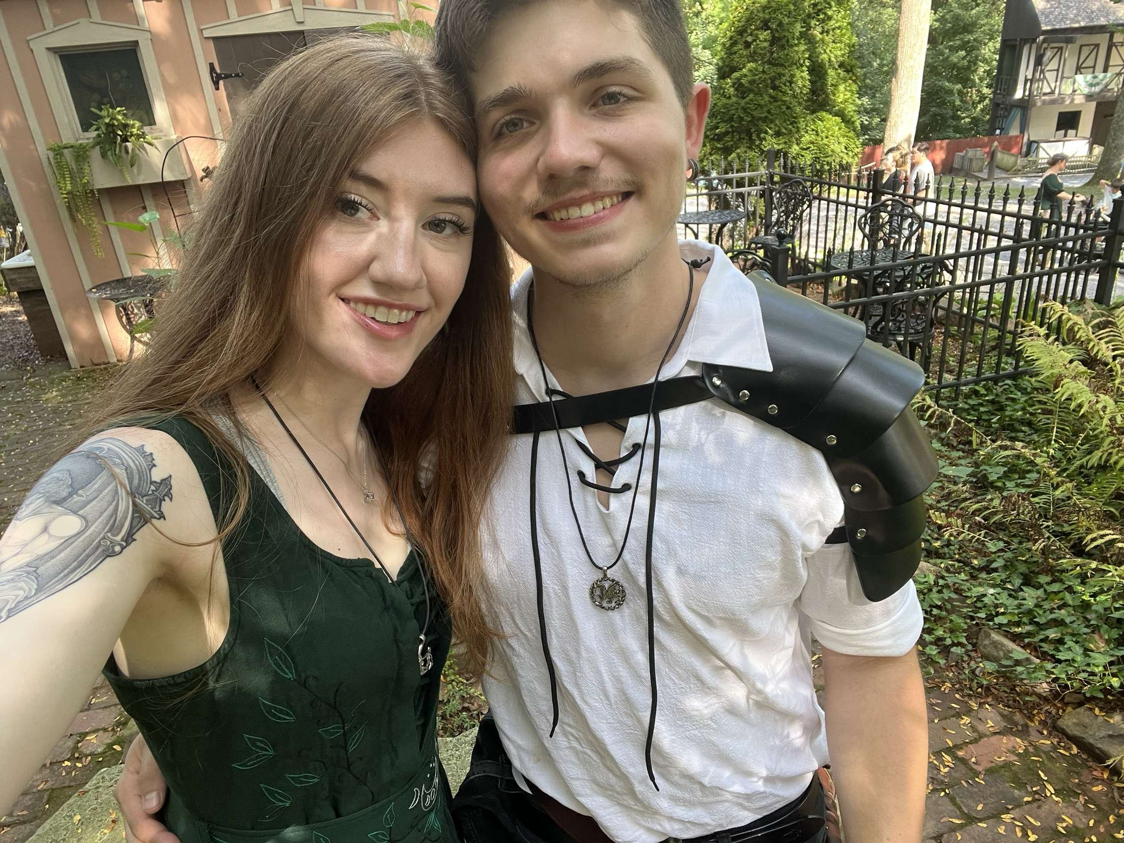A smiling young woman with long red hair and tattoos on her arm takes a selfie with a young man in a white shirt. They are outdoors in a garden or park with trees, plants, and a black iron fence. The man has short dark hair, wears earrings, a necklace, and holds a camera with a large lens. In the background, there are people near a pond and a house.