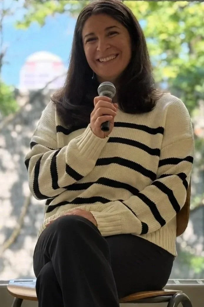 Smiling woman in a cream-and-black striped sweater speaking into a microphone outdoors