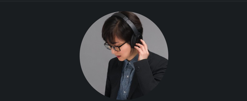 Woman with short dark hair, glasses, wearing a black blazer and gray shirt, listening to headphones.