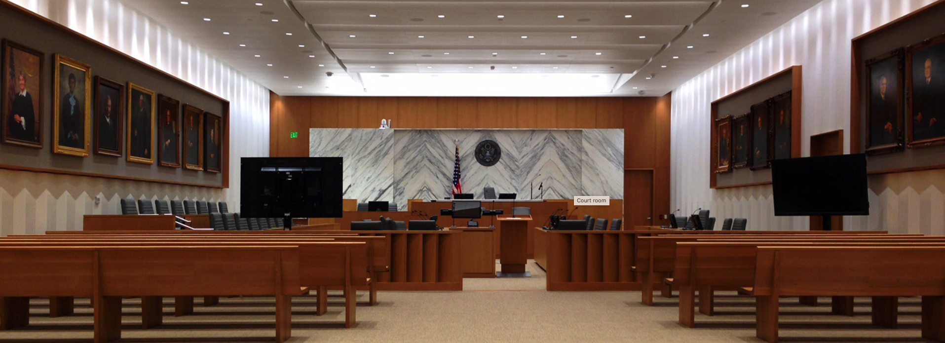 Empty court room with wooden benches, a marble wall backdrop with a seal, American flag, two large television screens, and portraits of judges or officials hanging on the walls.