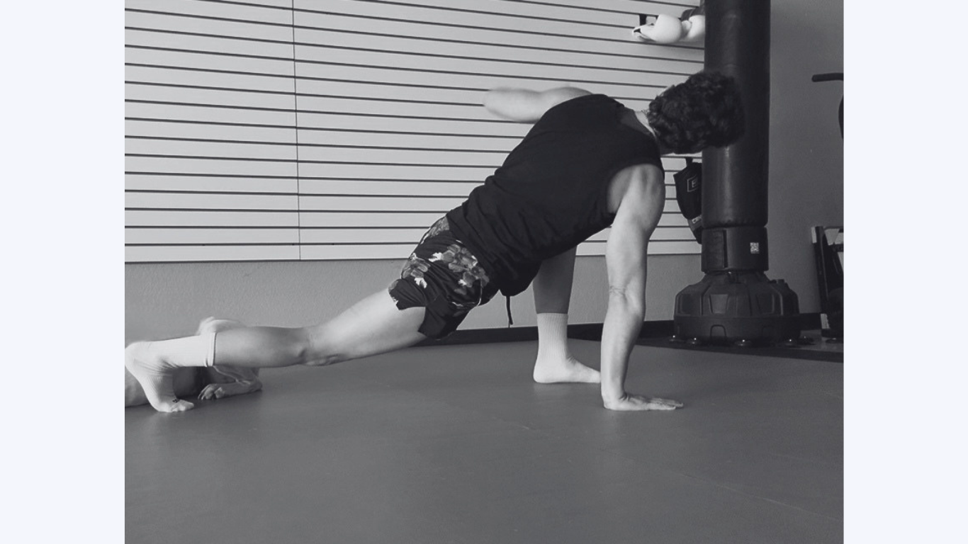 A man in workout clothes doing a one-arm push-up on the floor in a gym. His left hand is on the ground, legs extended, with a neutral background.