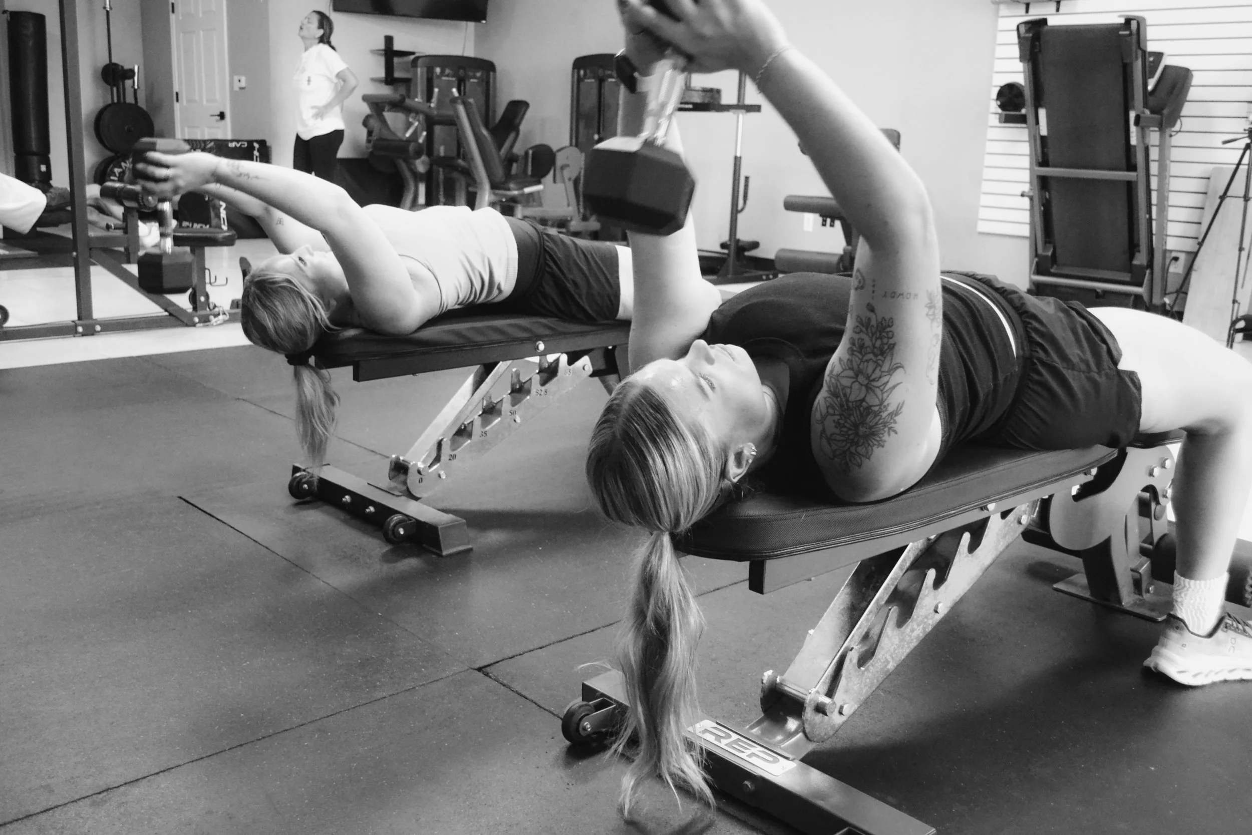 Two women lying on workout benches performing exercises with dumbbells in a gym. One woman is in the foreground, while the other is in the background. The gym has various exercise equipment and a television mounted on the wall.
