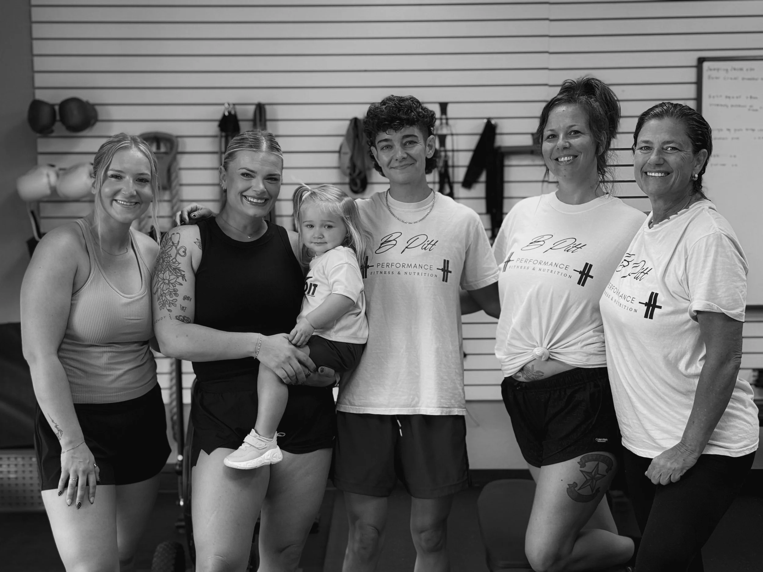 A group of six women and one young girl standing together in a fitness or sports facility, smiling at the camera. Some are wearing T-shirts with a logo that reads 'B Pitt Performance Fitness & Nutrition,' while others are in athletic clothing. The background features gym equipment and a whiteboard.