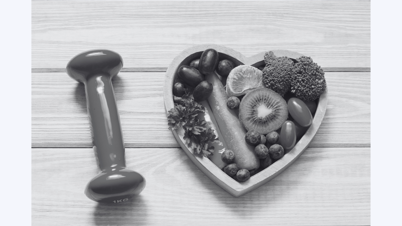 A heart-shaped bowl filled with various fruits and vegetables including berries, kiwi, broccoli, grapes, and a slice of citrus, placed on a wooden surface next to a 1 kg dumbbell.