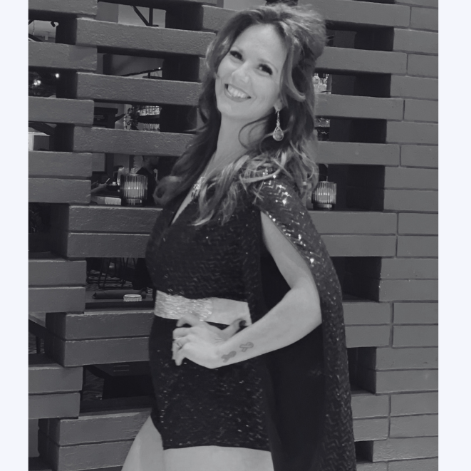 A woman in a sparkly dress and cape, smiling with her left hand on her hip, standing in front of a brick wall. It is a black-and-white photo.
