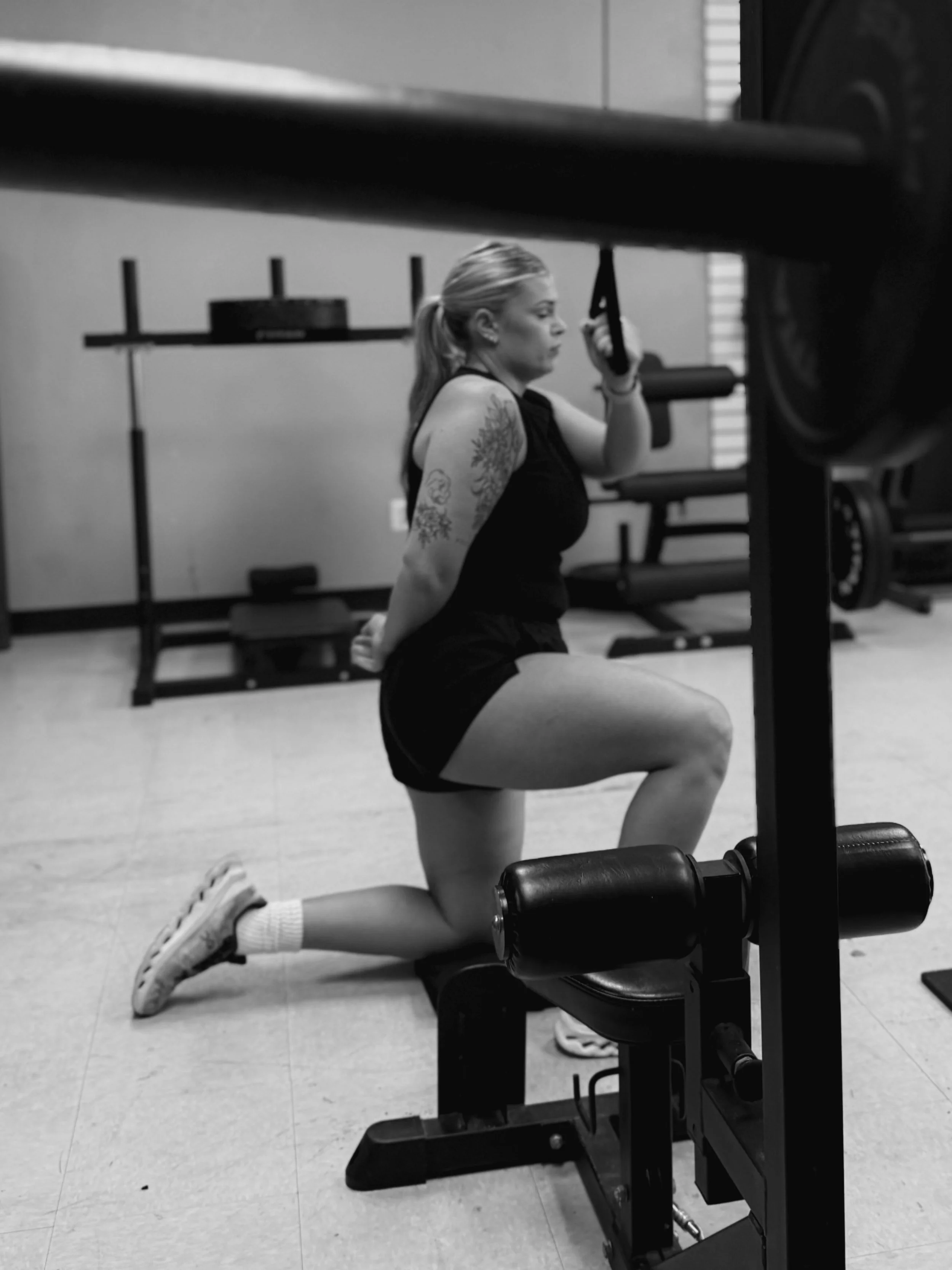 A woman with tattoos on her upper arm is performing a knee lunge exercise in a gym, using a workout bench and holding a strap with one hand while keeping her other hand behind her back.