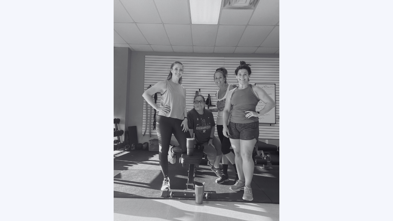Four women in workout attire posing inside a gym, with gym equipment and whiteboard in the background.