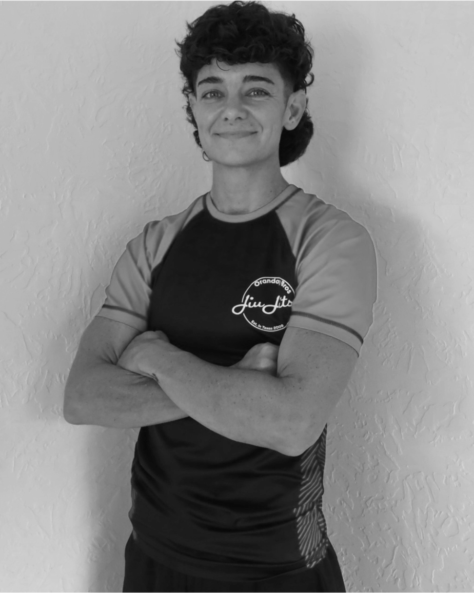 A young woman with short, curly hair and a confident smile, crossing her arms, wearing a sports T-shirt with a logo on the chest, standing in front of a textured wall.