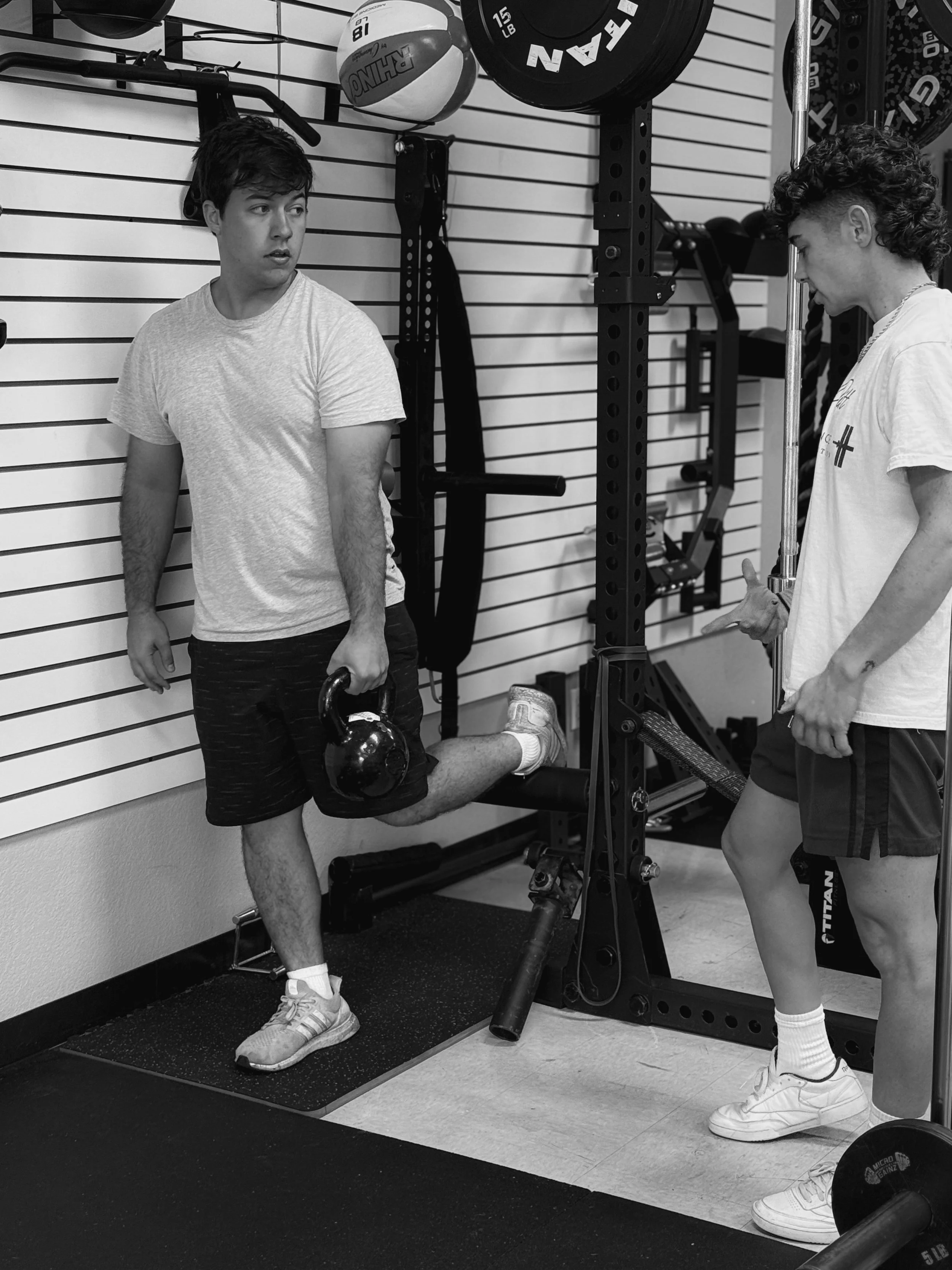 Coach and client are engaged in a workout session in a gym, one is holding a kettlebell with his left arm resting against the wall, and the other is standing nearby, giving instructions.