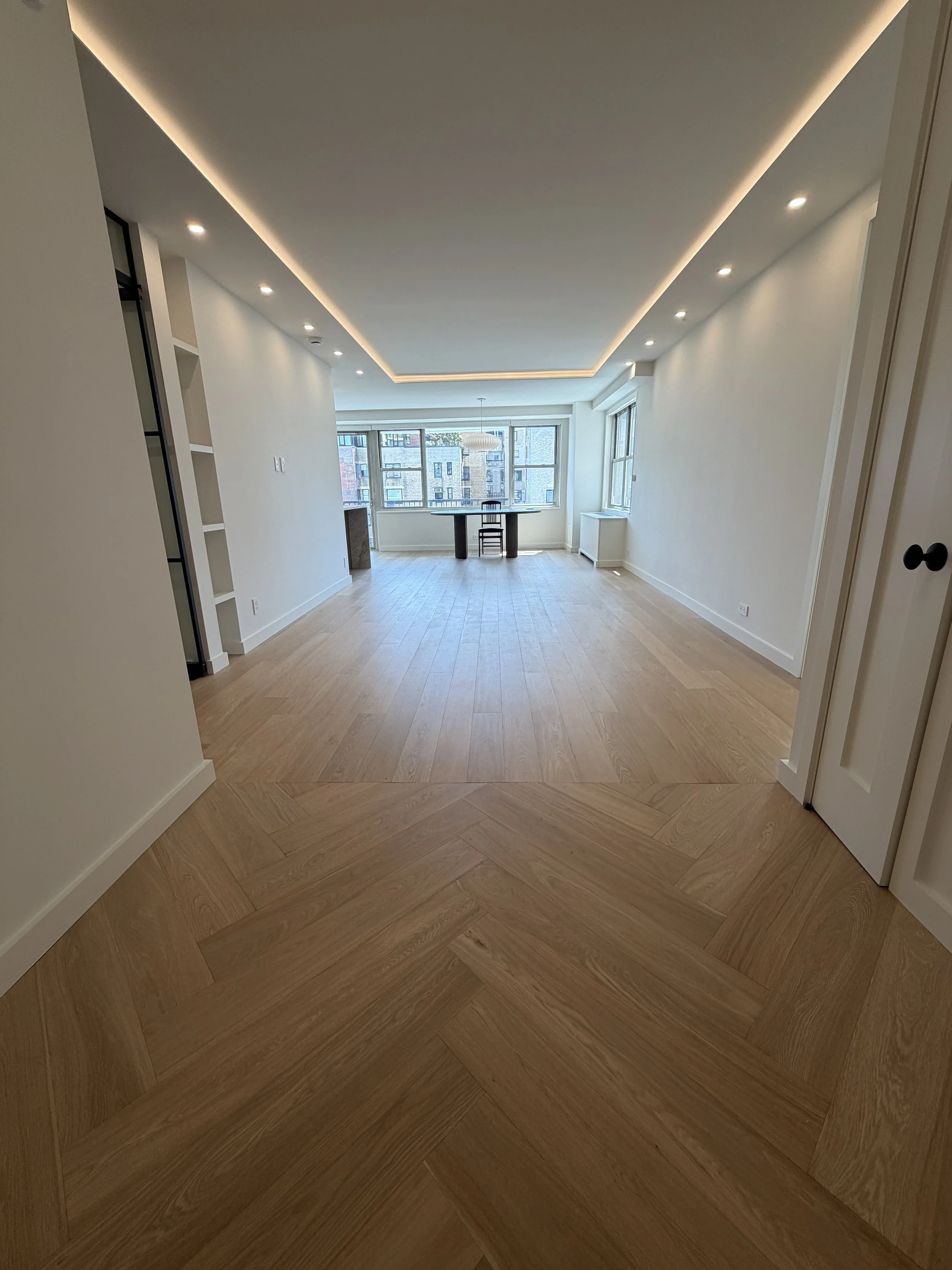 Empty living room with light wood floors, white walls, large windows, and a black table with a black chair in front.