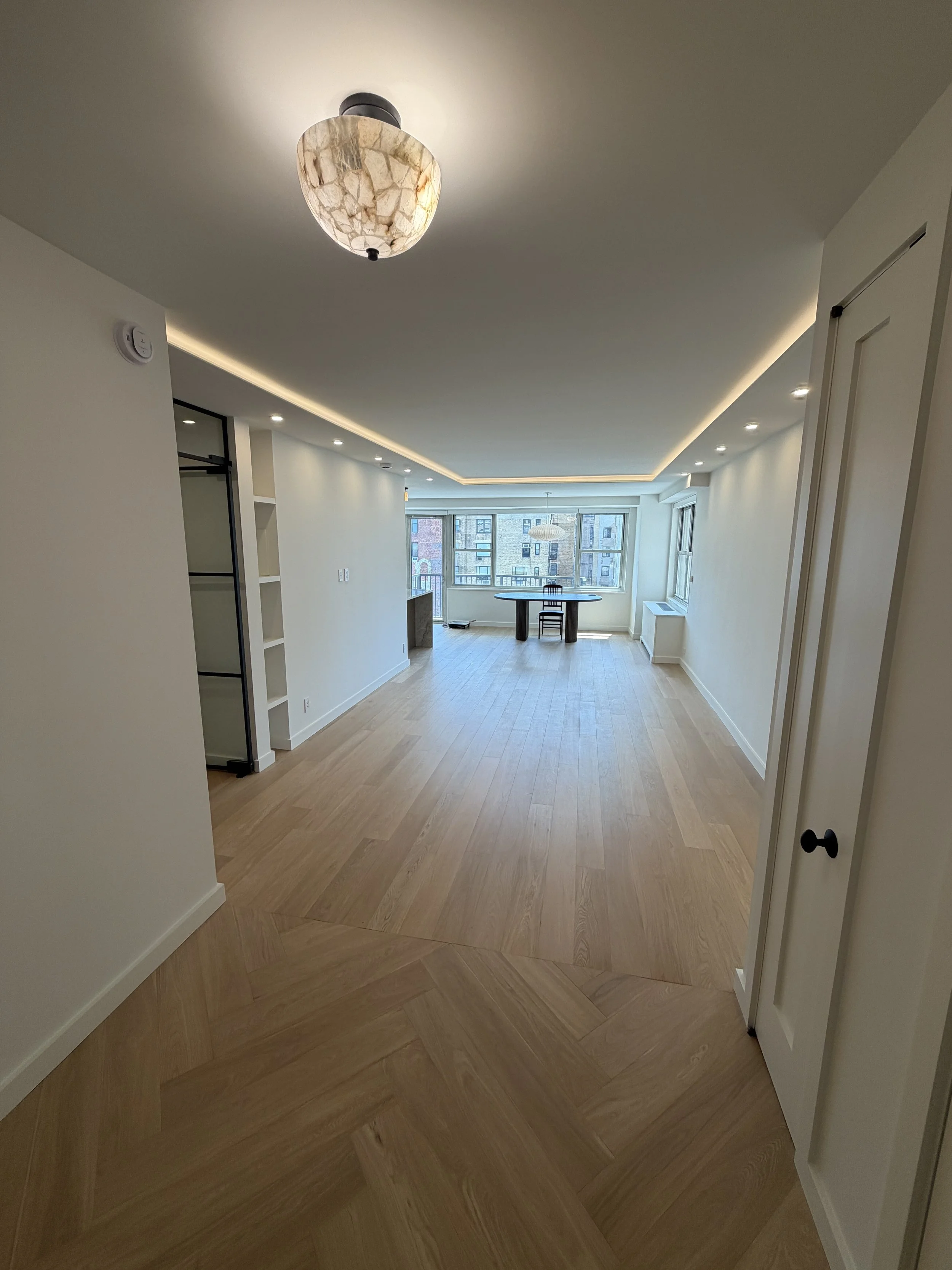 Interior of an apartment with light-colored hardwood floors, white walls, a round dining table, chairs, large windows, and ceiling lighting.