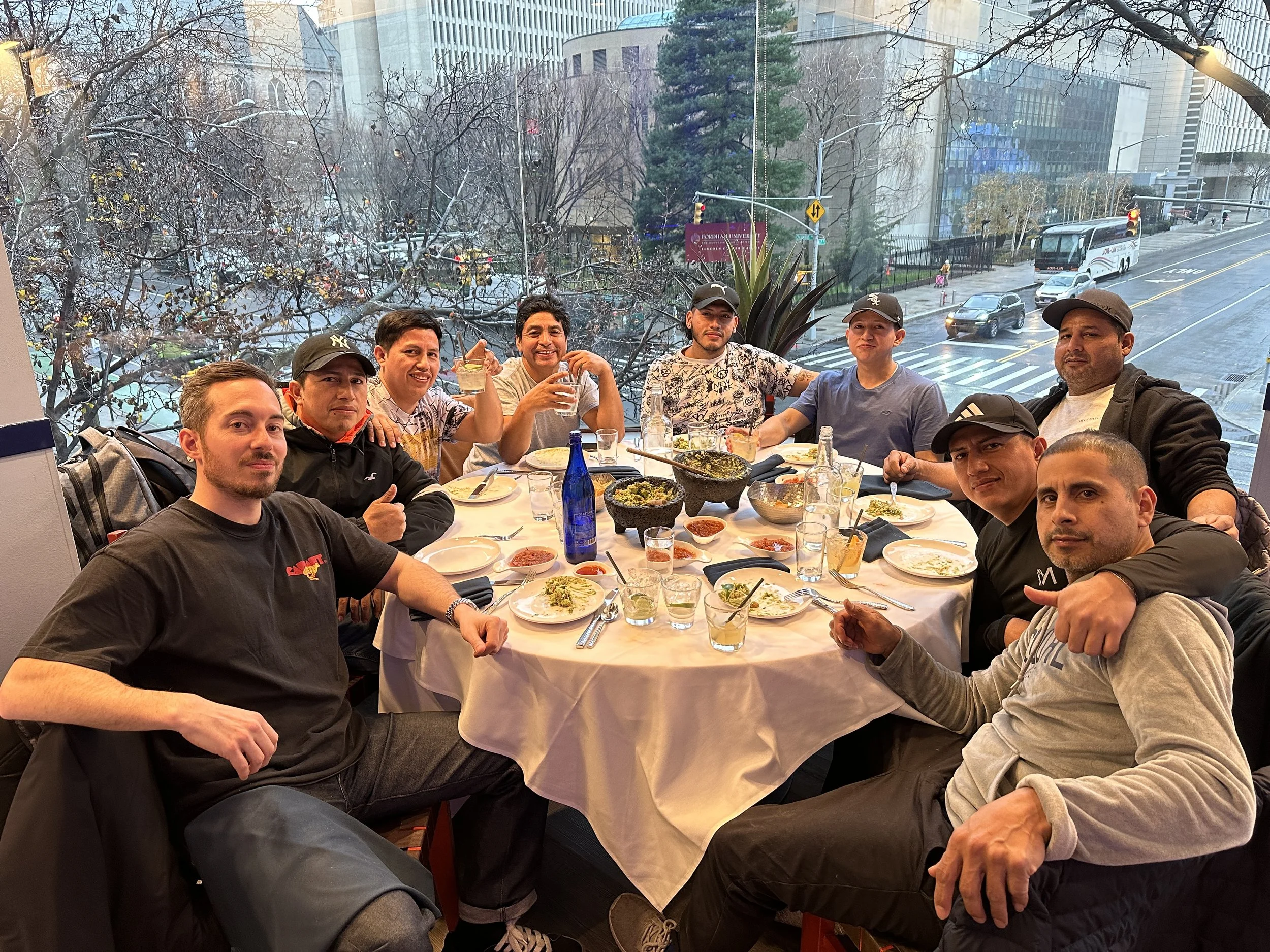 A group of men sitting around a table enjoying a meal indoors, with a large window behind them showing a city street with cars, a bus, trees, and buildings.