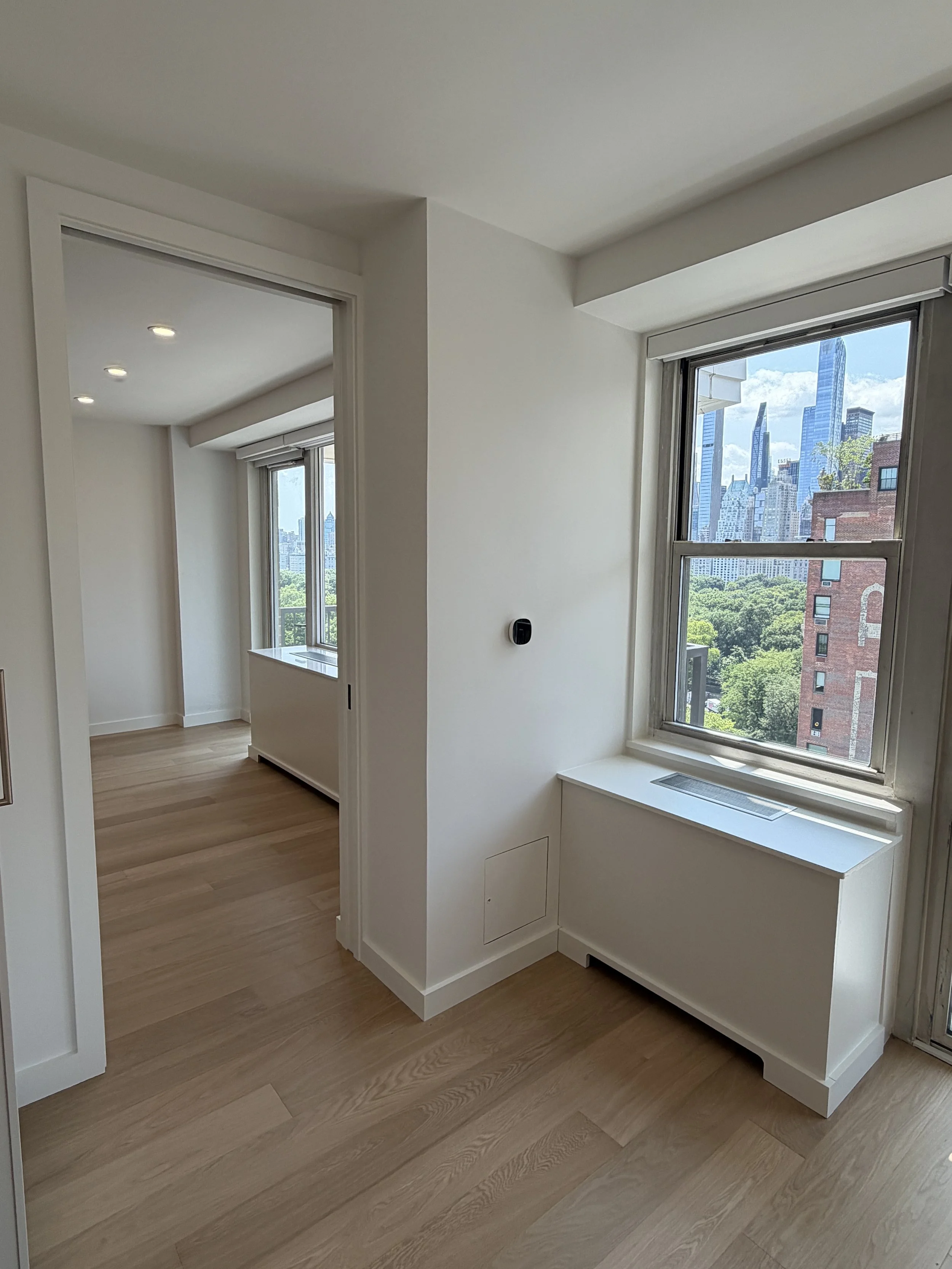 Interior view of a modern apartment with large windows showing a city skyline and green trees, hardwood floors, and white walls.