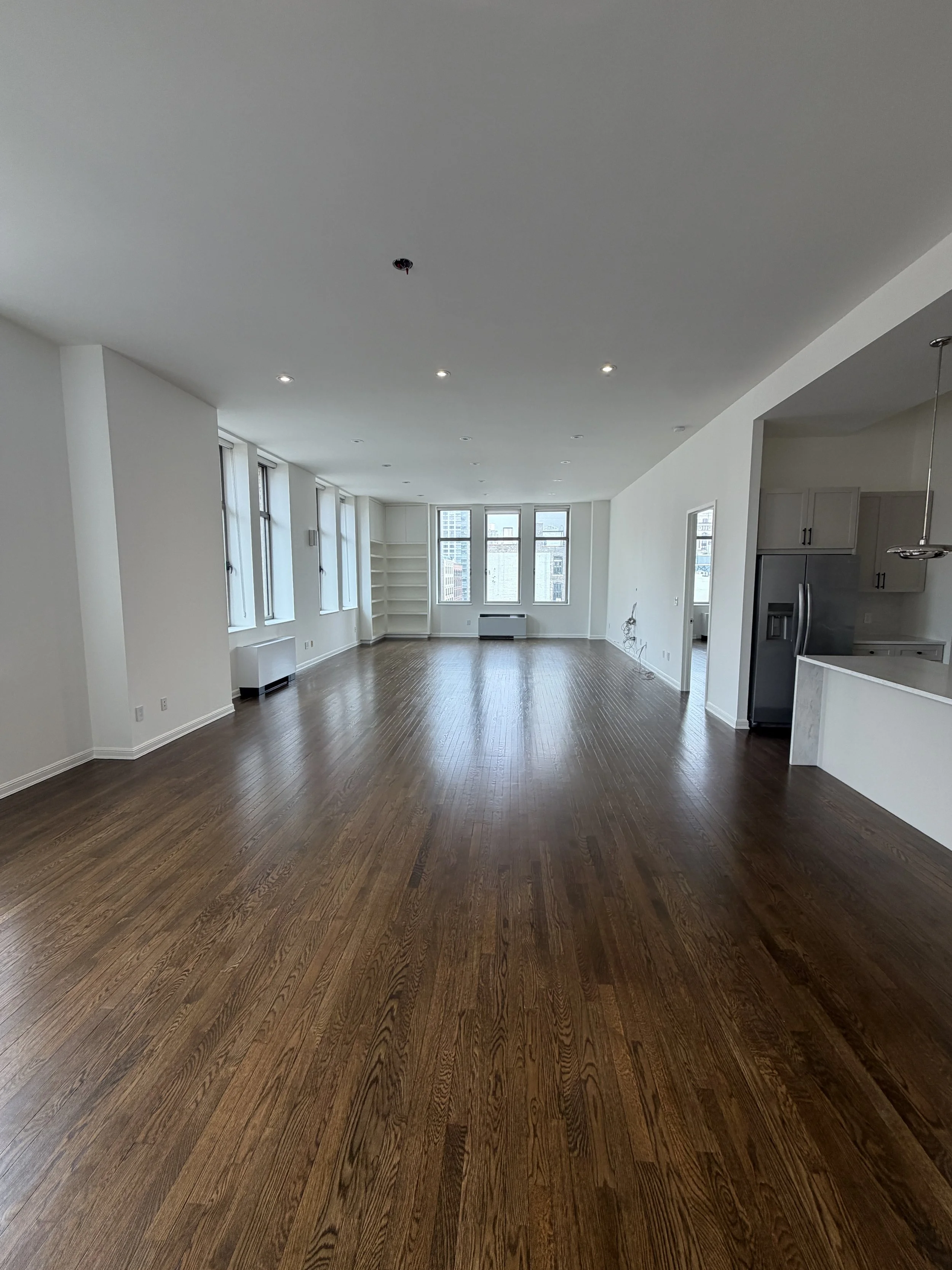 Empty living room with hardwood floors, multiple large windows, and a view of city buildings outside. The ceiling has recessed lighting, and there is an open kitchen area visible to the right.
