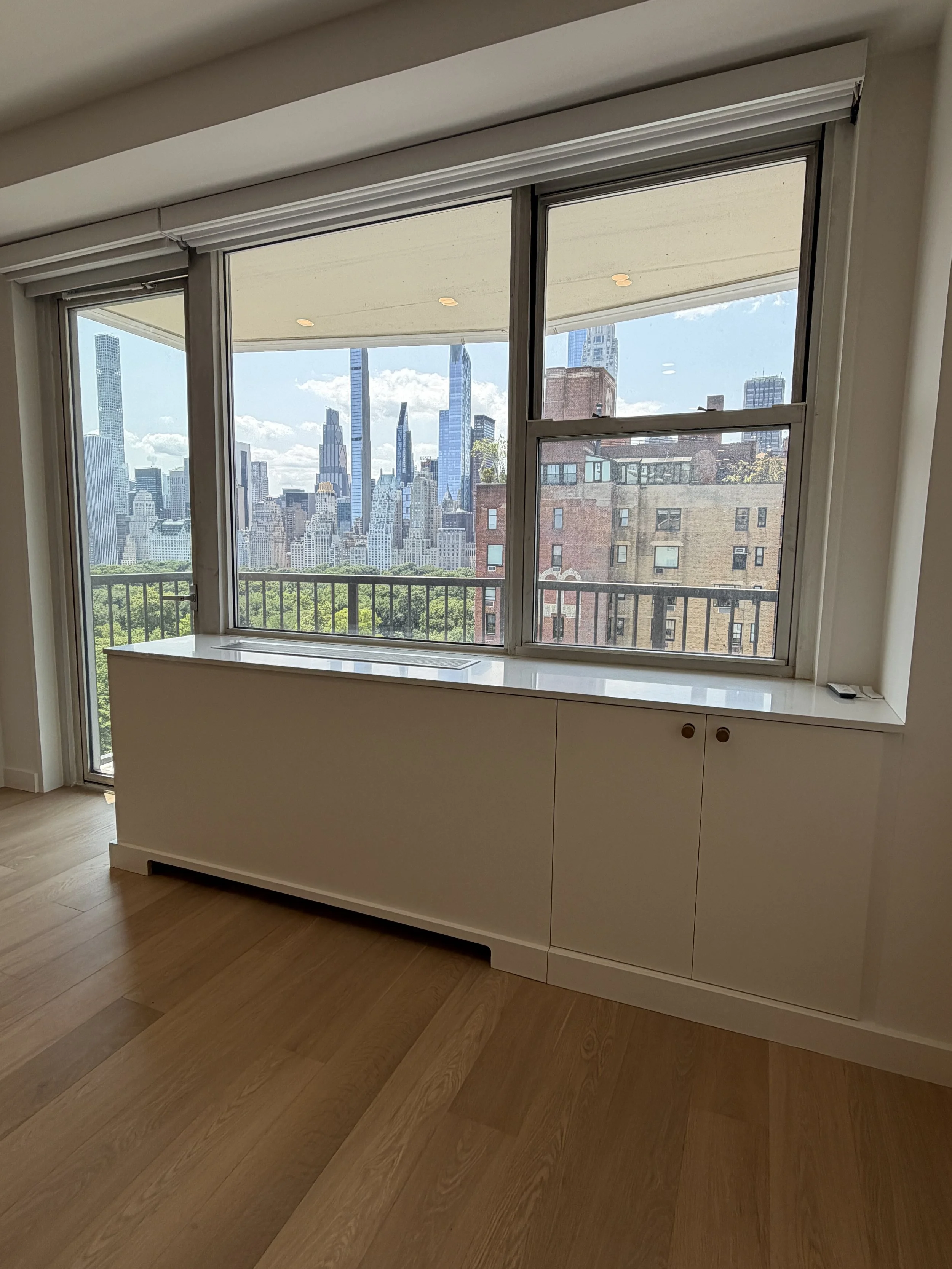 Interior view of a room with large windows displaying a city skyline, hardwood floors, and a white built-in cabinet.